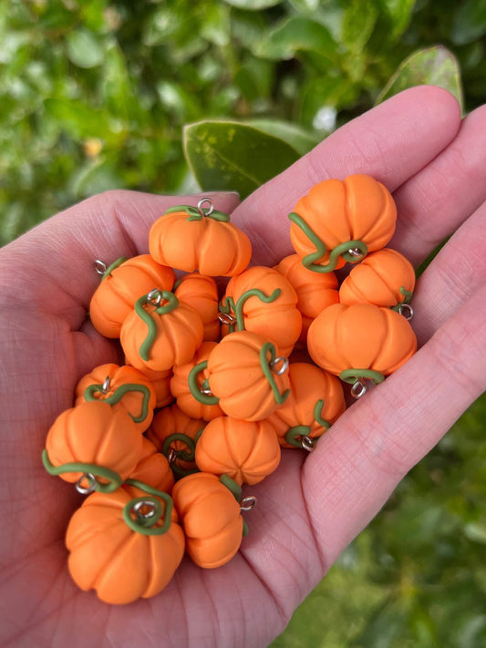 A hand is showcasing a set of Pumpkin Polymer Clay Earrings, featuring small, orange pumpkins with green stems. The lush green foliage in the background accentuates their unique, handcrafted design.