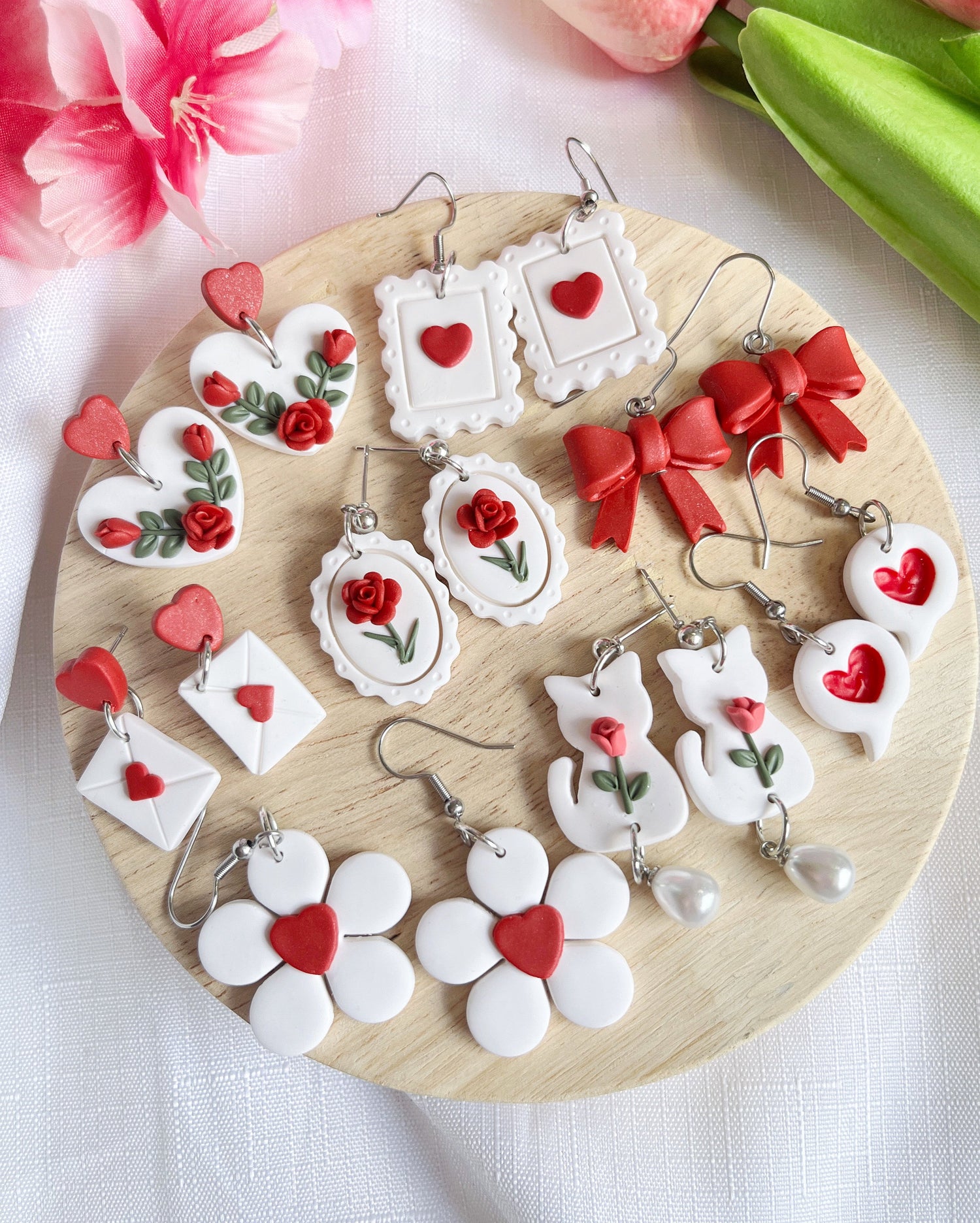 A round wooden board displays various handmade earrings, featuring designs with hearts, roses, envelopes, bows, and abstract shapes. Predominantly red and white, with a few earrings incorporating pearl-like elements.