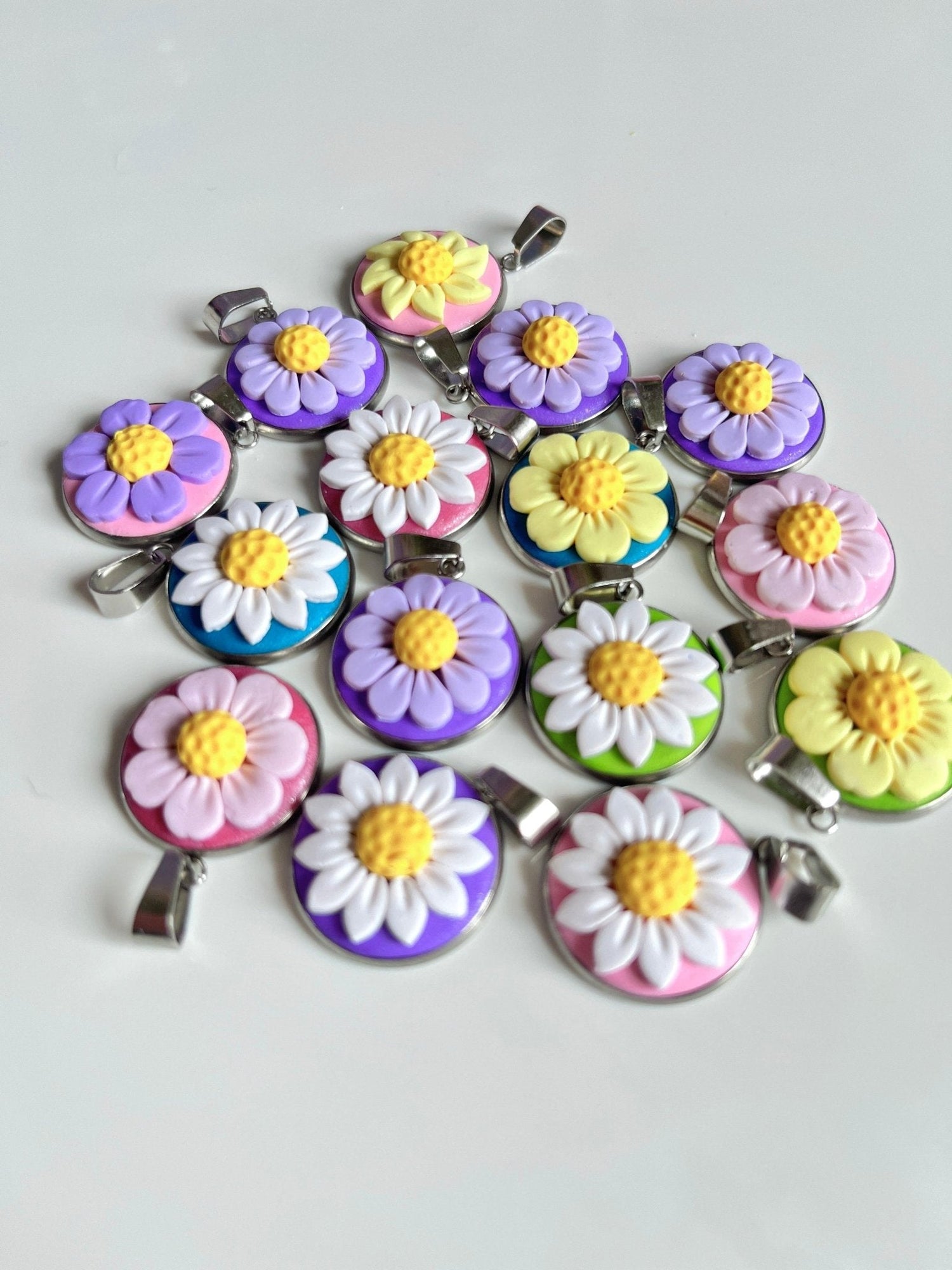 Assorted colorful daisy charms with yellow centers, featuring purple, pink, and green backgrounds, arranged on a white surface. Each charm has a metal loop for attaching.