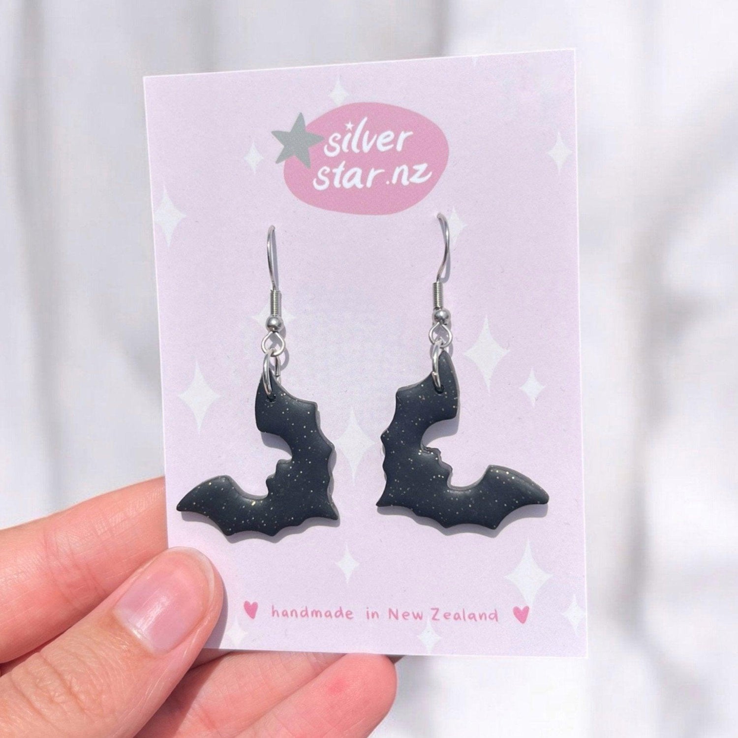 Close-up of a hand holding a card with two earrings shaped like bats. The earrings are black with a subtle sparkle. The card is pink with silver star.nz and handmade in New Zealand printed on it.