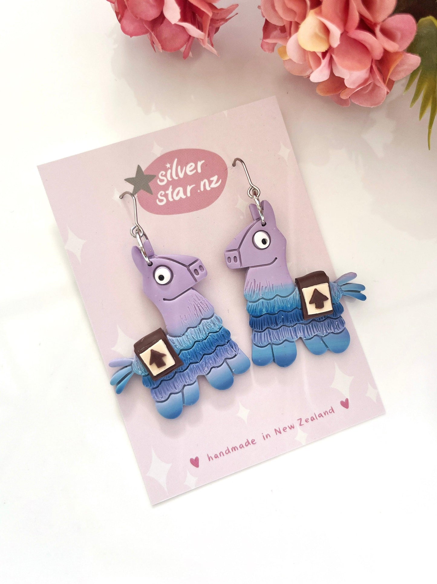 The Fortnite Llama Polymer Clay Earrings are a vibrant pair shaped like llamas, crafted from polymer clay in delightful shades of purple and blue, featuring a small pouch design. These handmade earrings come with stainless steel hooks and are elegantly presented on a card labeled "silver star.nz," with pink flowers accentuating the background.