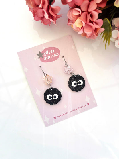 A pair of whimsical Soot Sprite earrings, crafted from polymer clay and featuring googly eyes and pink bead accents, are displayed on a pink silver star NZ card. Surrounded by pink flowers, these handmade treasures from New Zealand offer a charming, unique design.