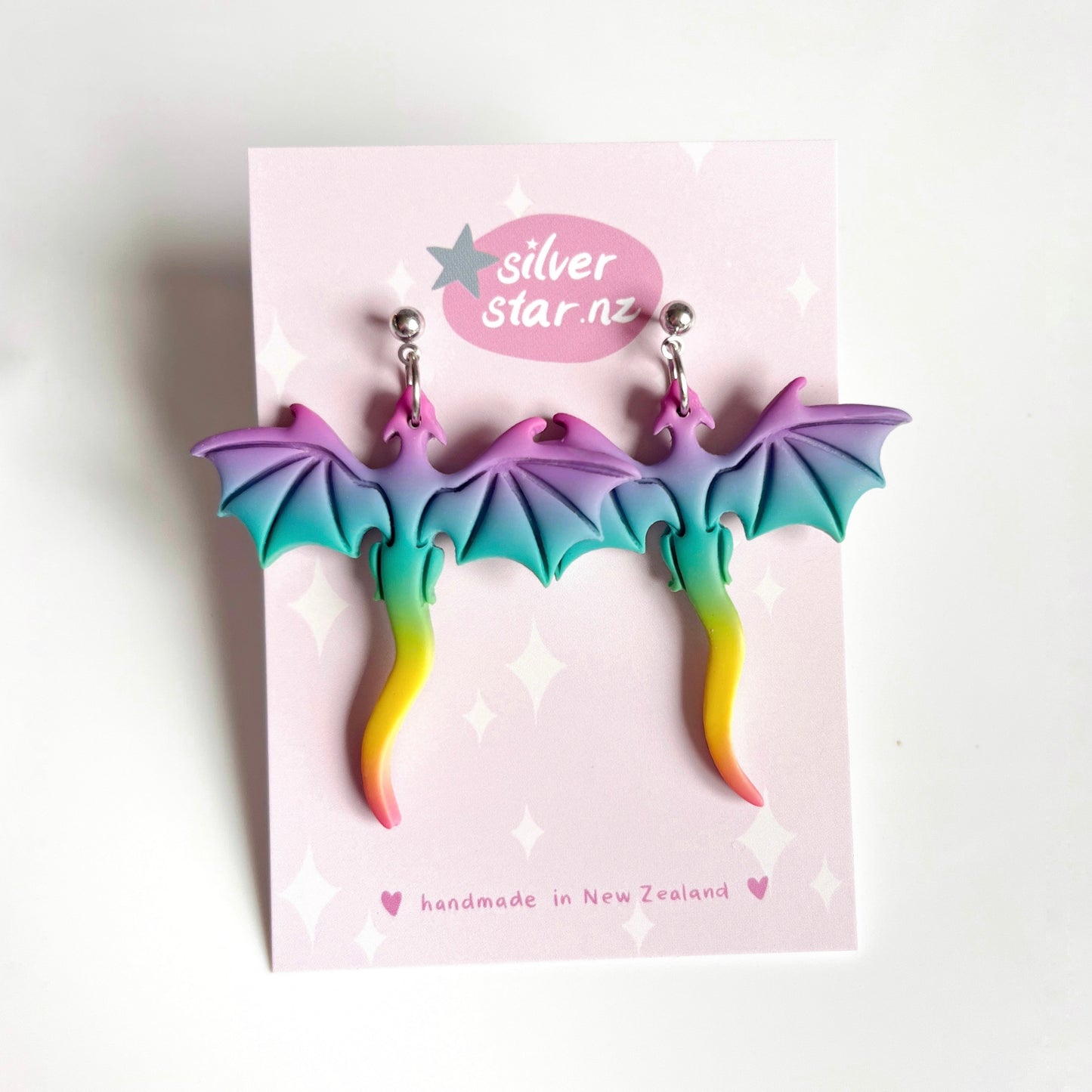 The Rainbow Dragon Polymer Clay Earrings by silverstar.nz boast a vibrant purple-to-blue-to-yellow gradient design, with silver stud attachments. They hang on a pink card adorned with white stars.