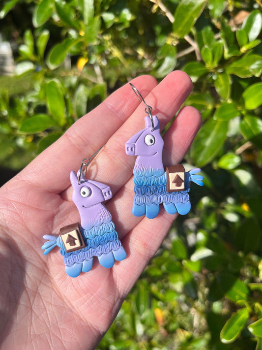 A close-up shot captures a hand holding vibrant Fortnite Llama Polymer Clay Earrings, featuring llamas crafted in purple and blue shades with intricate tiny saddlebags. Beneath lush green leaves shining in the bright sunlight, the earrings dangle from stainless steel hooks.