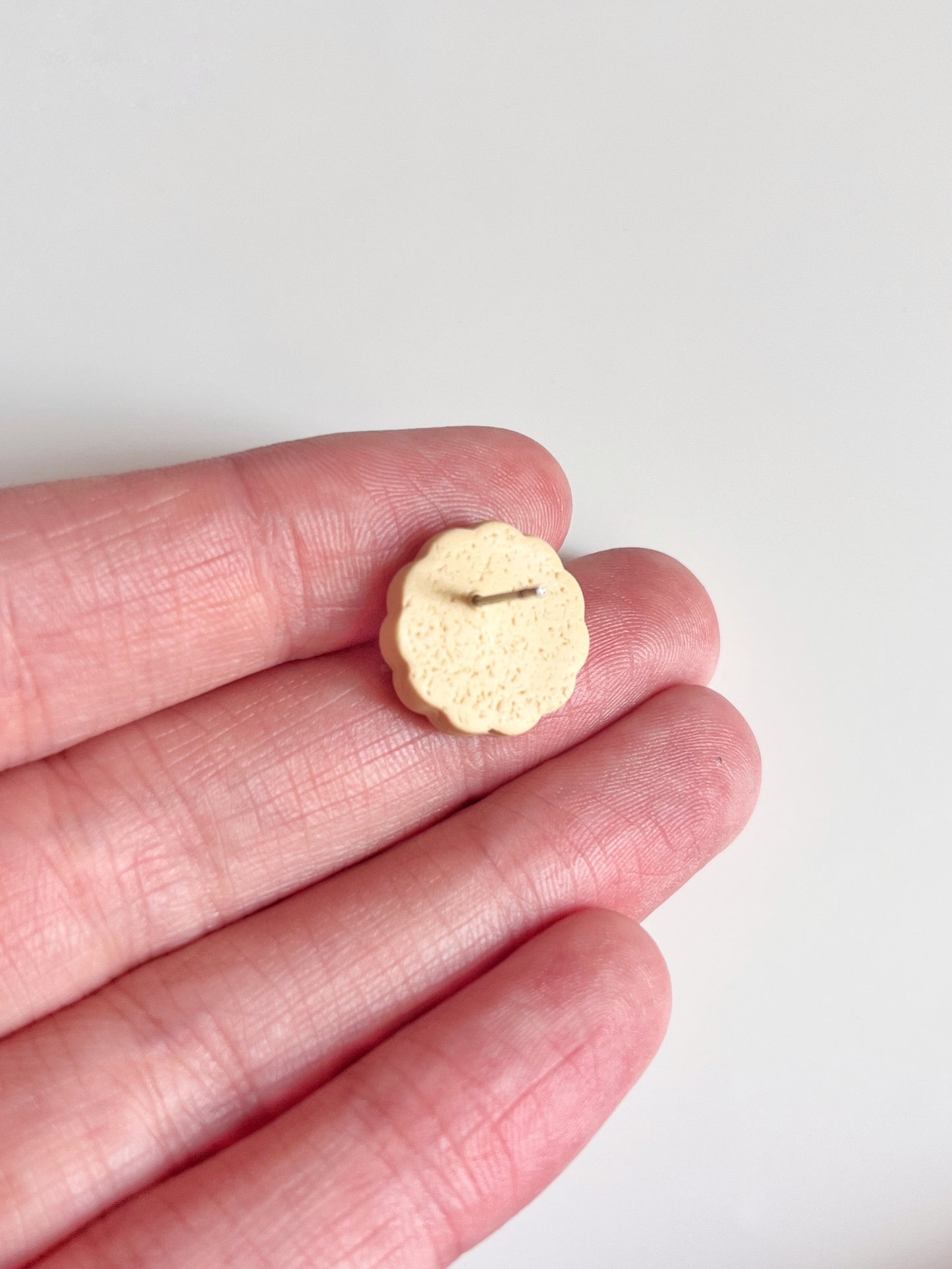 A small, round object resembling a cookie rests on three fingers against a white background. Crafted with polymer clay, these earrings from silverstar.nz feature a scalloped edge and an indented pattern, revealing the unique design of the Hundreds and Thousands Biscuit Stud Earrings.