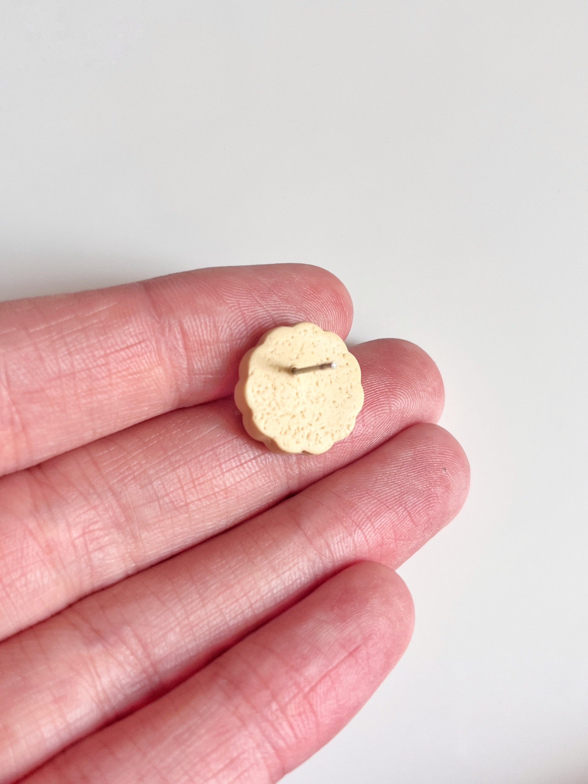 A small, round object resembling a cookie rests on three fingers against a white background. Crafted with polymer clay, these earrings from silverstar.nz feature a scalloped edge and an indented pattern, revealing the unique design of the Hundreds and Thousands Biscuit Stud Earrings.