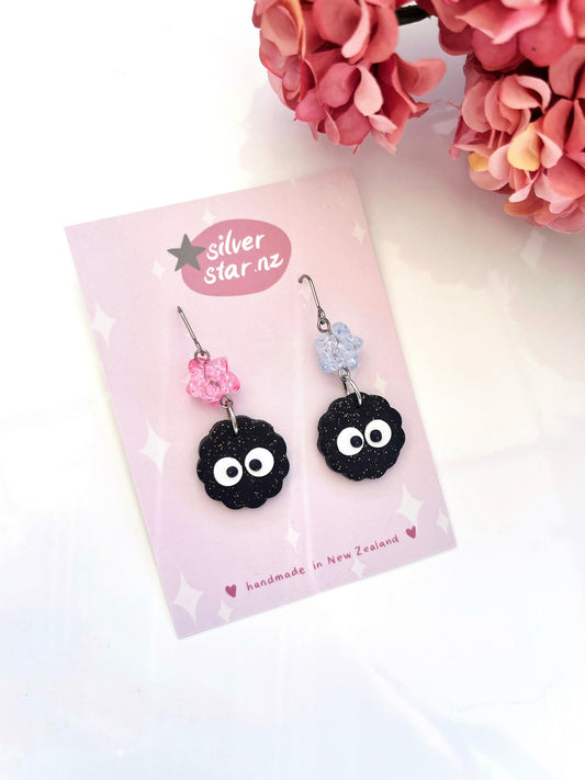 The Soot Sprite Polymer Clay Earrings with Bead boast a unique design featuring charming black soot sprite charms with googly eyes, complemented by pink and purple flower beads. These earrings are made from polymer clay and are presented on a silver card adorned with the silver star.nz logo, set against a backdrop of pink hydrangea flowers.