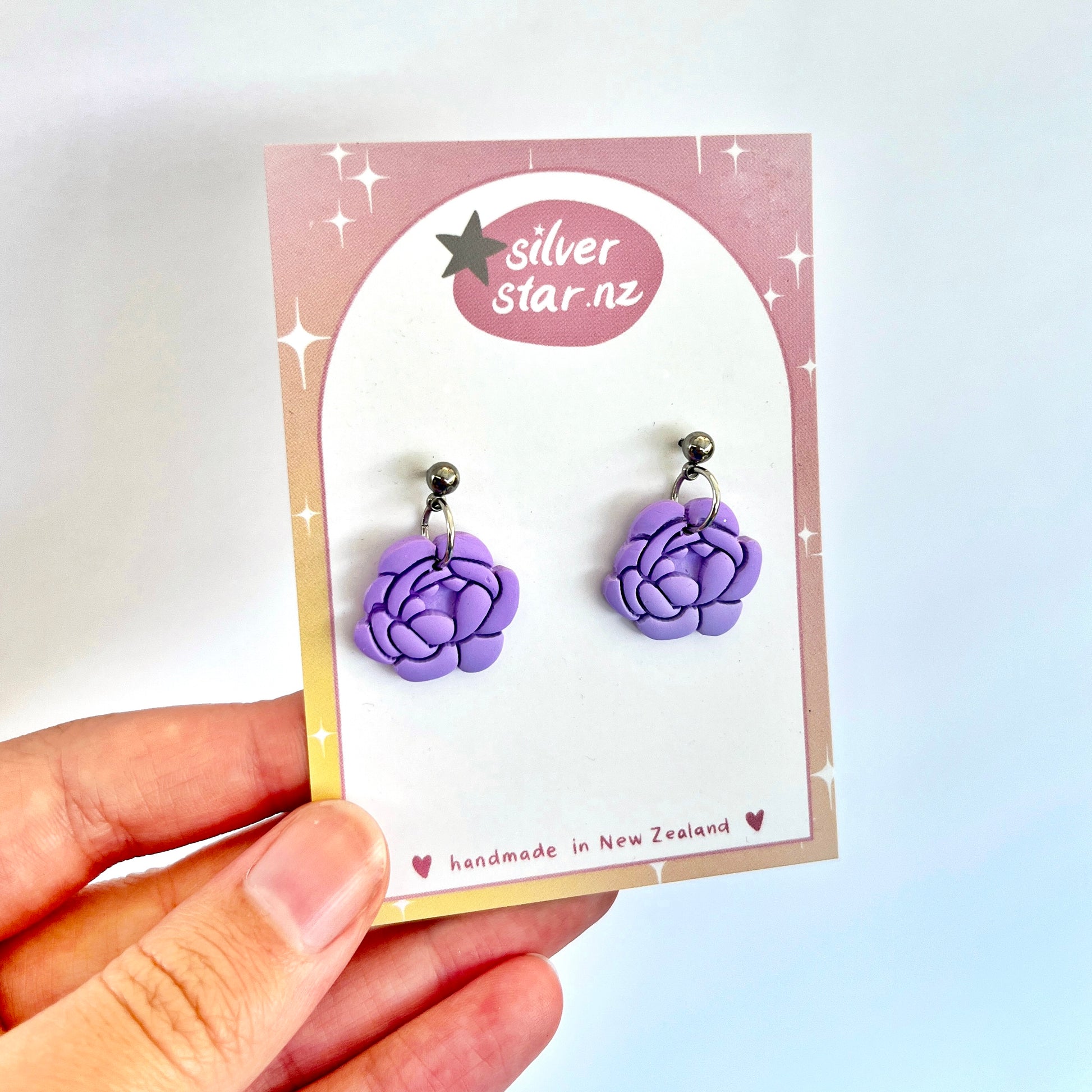 A hand displays a pair of Peony Polymer Clay Earrings, uniquely designed and made from polymer clay. The card showcasing these earrings features a pink and white sparkle motif with the text "silverstar.nz" and "handmade in New Zealand.