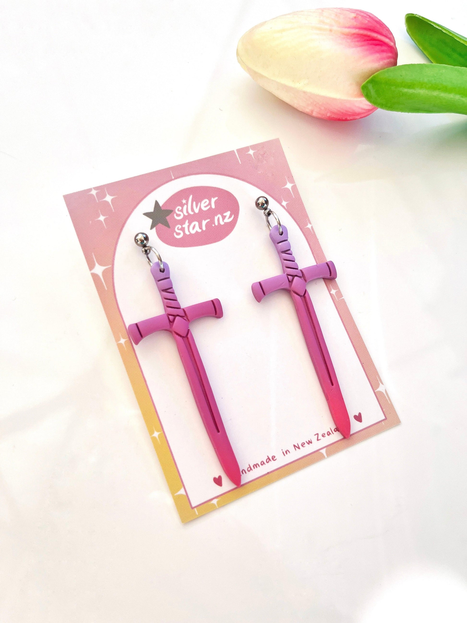 A pair of Sword Polymer Clay Earrings, elegantly displayed on a card featuring the silver star.nz logo, showcases a gradient background adorned with star patterns. Nearby, a pink and white tulip with vibrant green leaves adds to the composition.