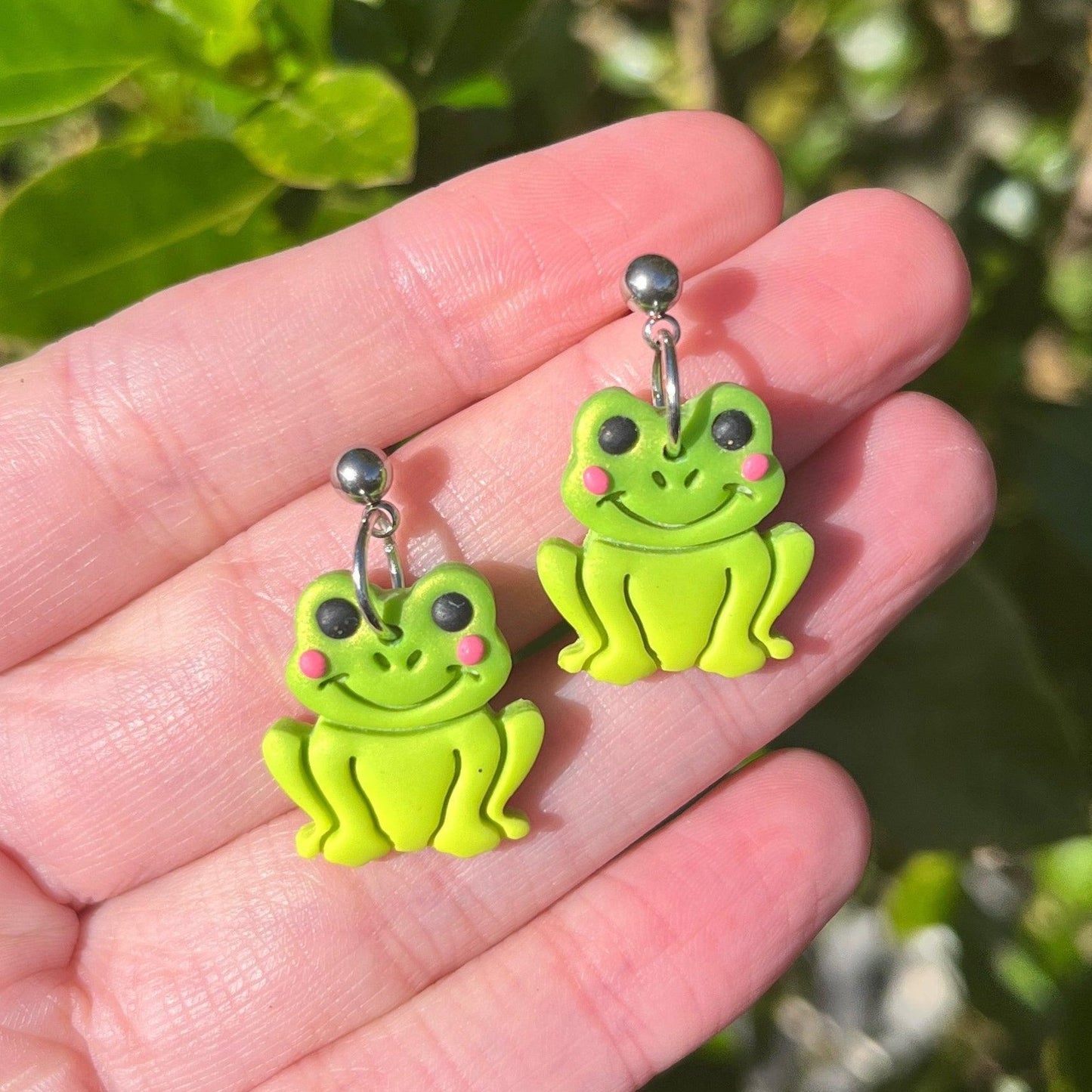 A hand displays a charming pair of Frog Polymer Clay Earrings, designed as playful green frog faces with cute pink cheeks and black eyes. Gleaming silver studs adorn each earring, set against a backdrop of vibrant green leaves basking in bright sunlight. These handmade earrings embody the whimsical essence of nature.