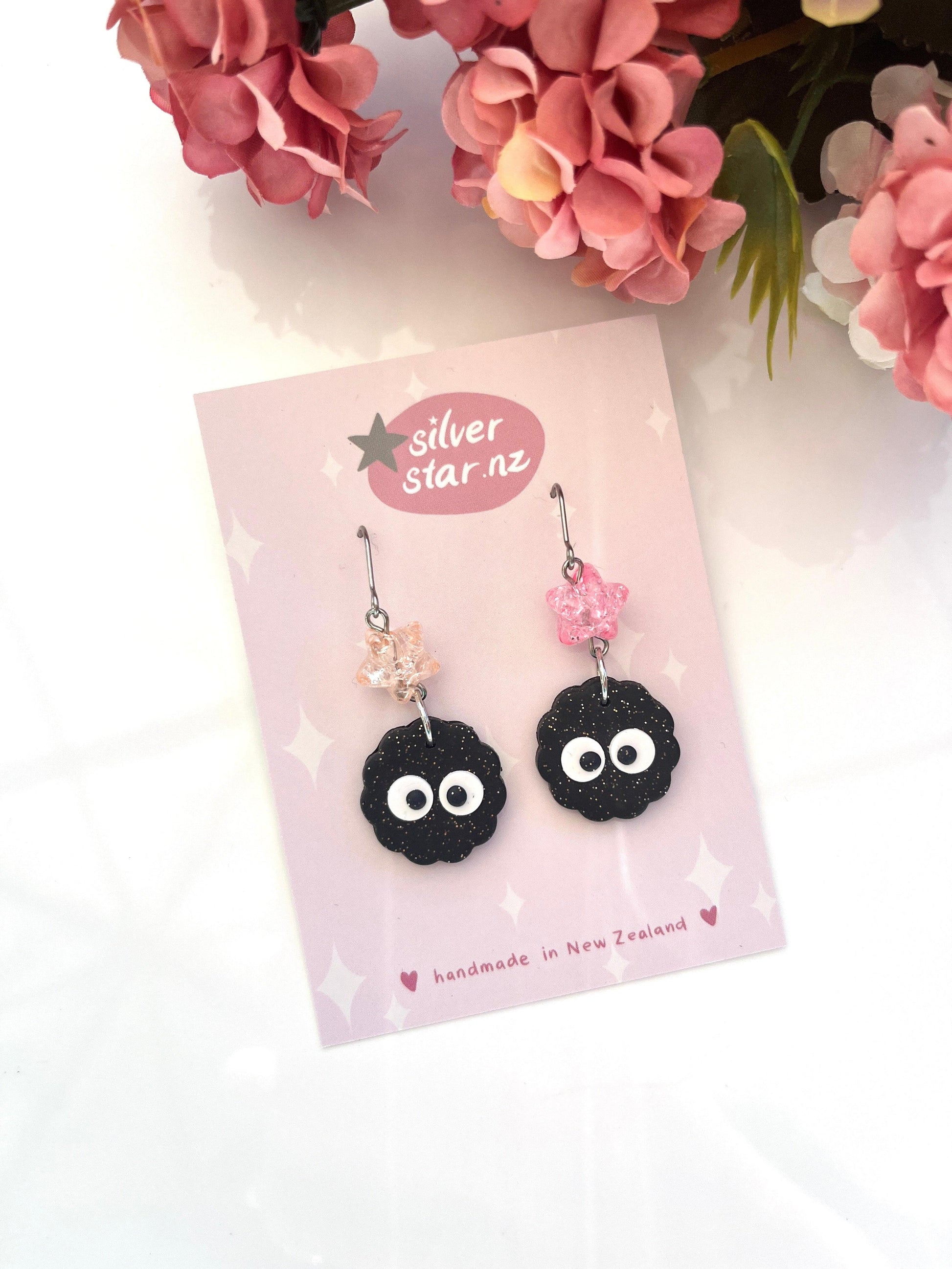 These expertly crafted Soot Sprite Polymer Clay Earrings with Bead from silver star.nz feature a unique design. The earrings display black flower shapes with googly eyes and pink beaded tops, surrounded by pink flowers, and are handmade in New Zealand.