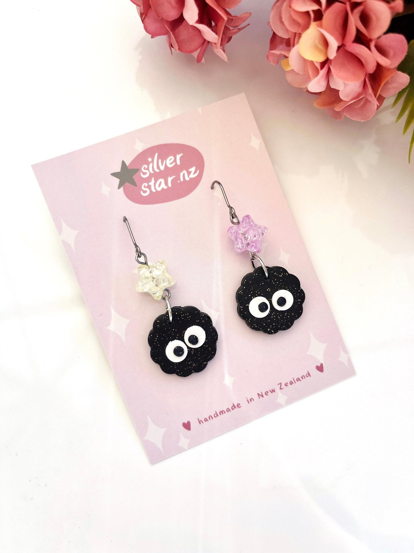 The Soot Sprite Polymer Clay Earrings with Bead are a distinctive pair of dangling earrings, featuring black, sparkling round charms with white eyes. They are attached to floral hooks and displayed on a pink card that reads "Handmade in New Zealand" and "silver star.nz," complemented by decorative pink flowers in the background.