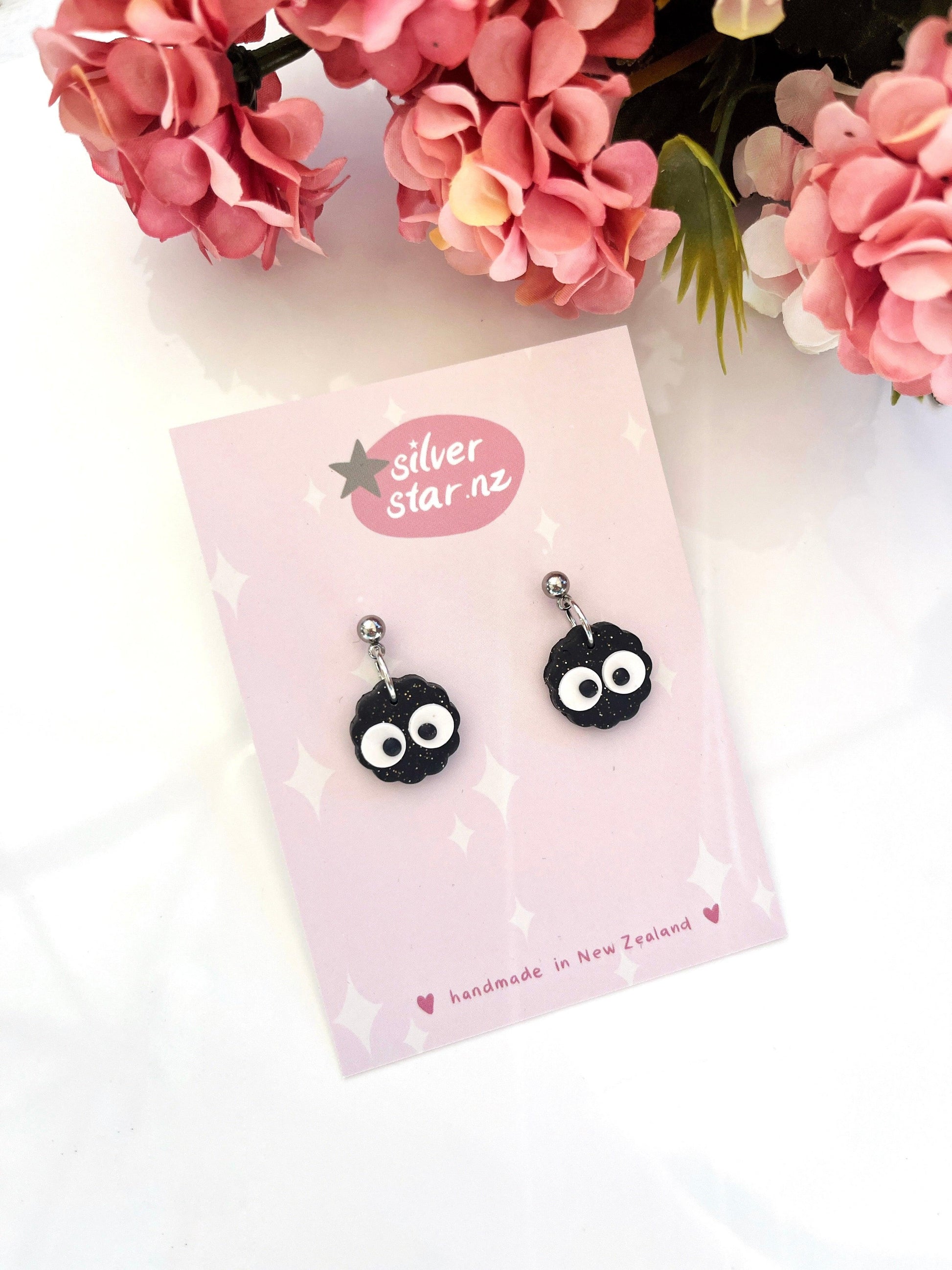 The Mini Soot Sprite Polymer Clay Earrings feature a unique design with black polymer clay adorned with white googly eyes and pink flowers. These handmade earrings are set on a pink card labeled silver star.nz, and they come with stainless steel hooks, showcasing craftsmanship from New Zealand.