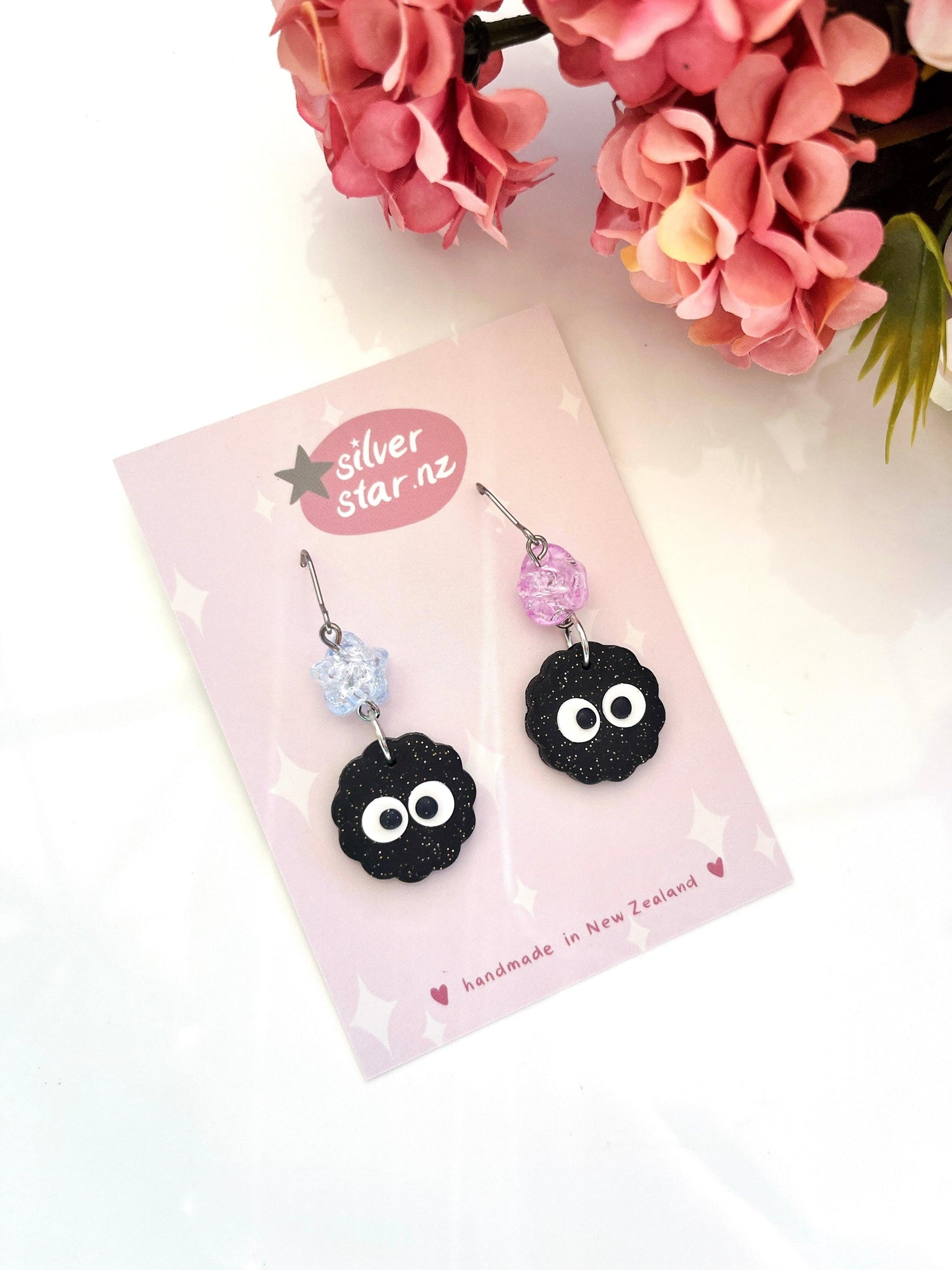 These Soot Sprite Polymer Clay Earrings with Beads are handmade and come on a card labeled Silver Star NZ. They showcase a unique design featuring adorable black soot sprite faces with big eyes, complemented by pastel beads—one in blue and the other in pink. Pink flowers provide a charming backdrop.