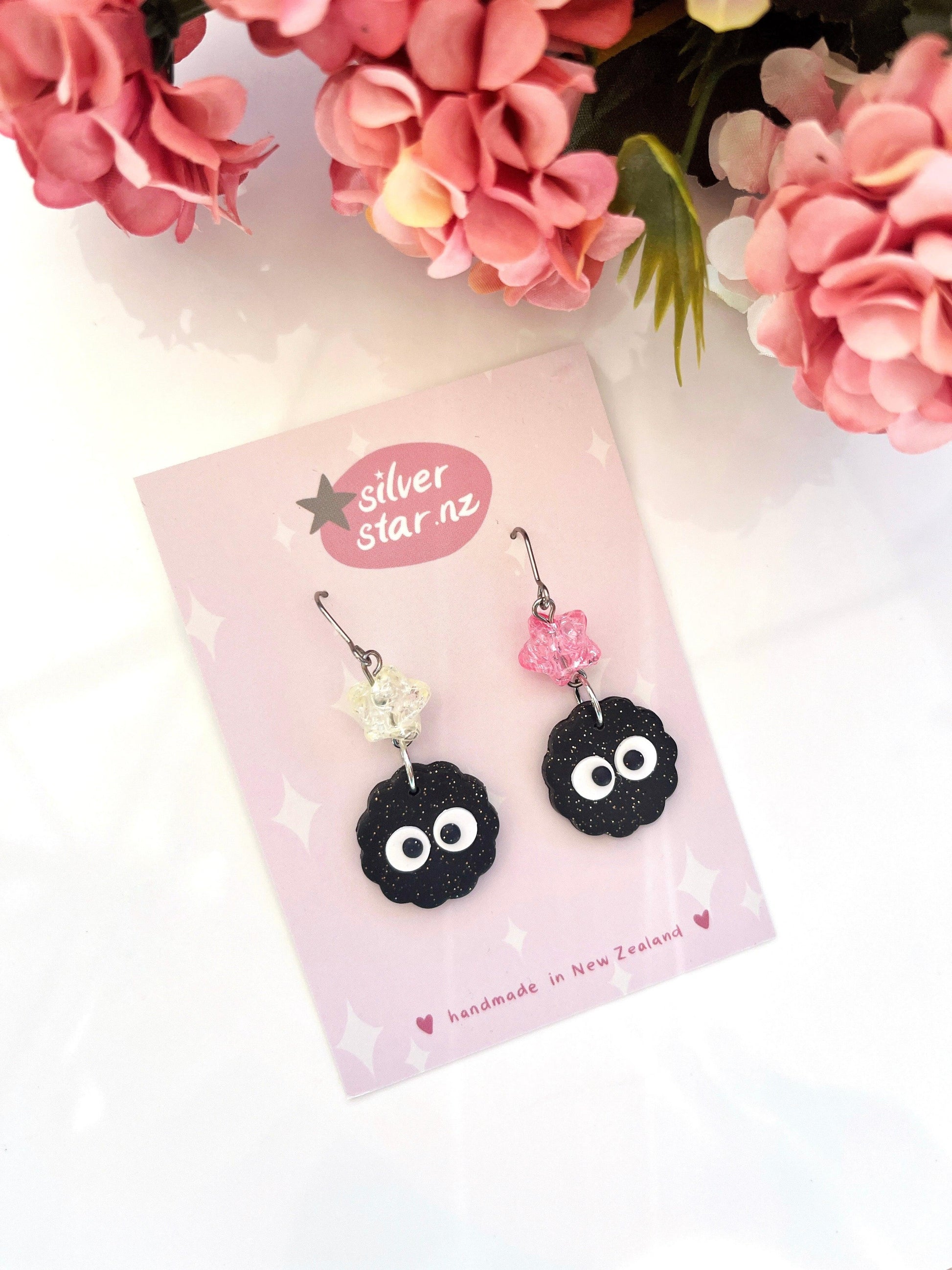 The Soot Sprite Polymer Clay Earrings with Bead are a pair of handcrafted earrings showcasing distinct black cloud-like forms and dangling beads in clear and pink hues. These unique earrings come displayed on a pink card labeled Silver Star NZ, enhanced by floral decorations.