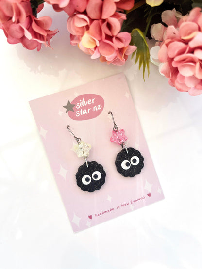 The Soot Sprite Polymer Clay Earrings with Bead are a pair of handcrafted earrings showcasing distinct black cloud-like forms and dangling beads in clear and pink hues. These unique earrings come displayed on a pink card labeled Silver Star NZ, enhanced by floral decorations.