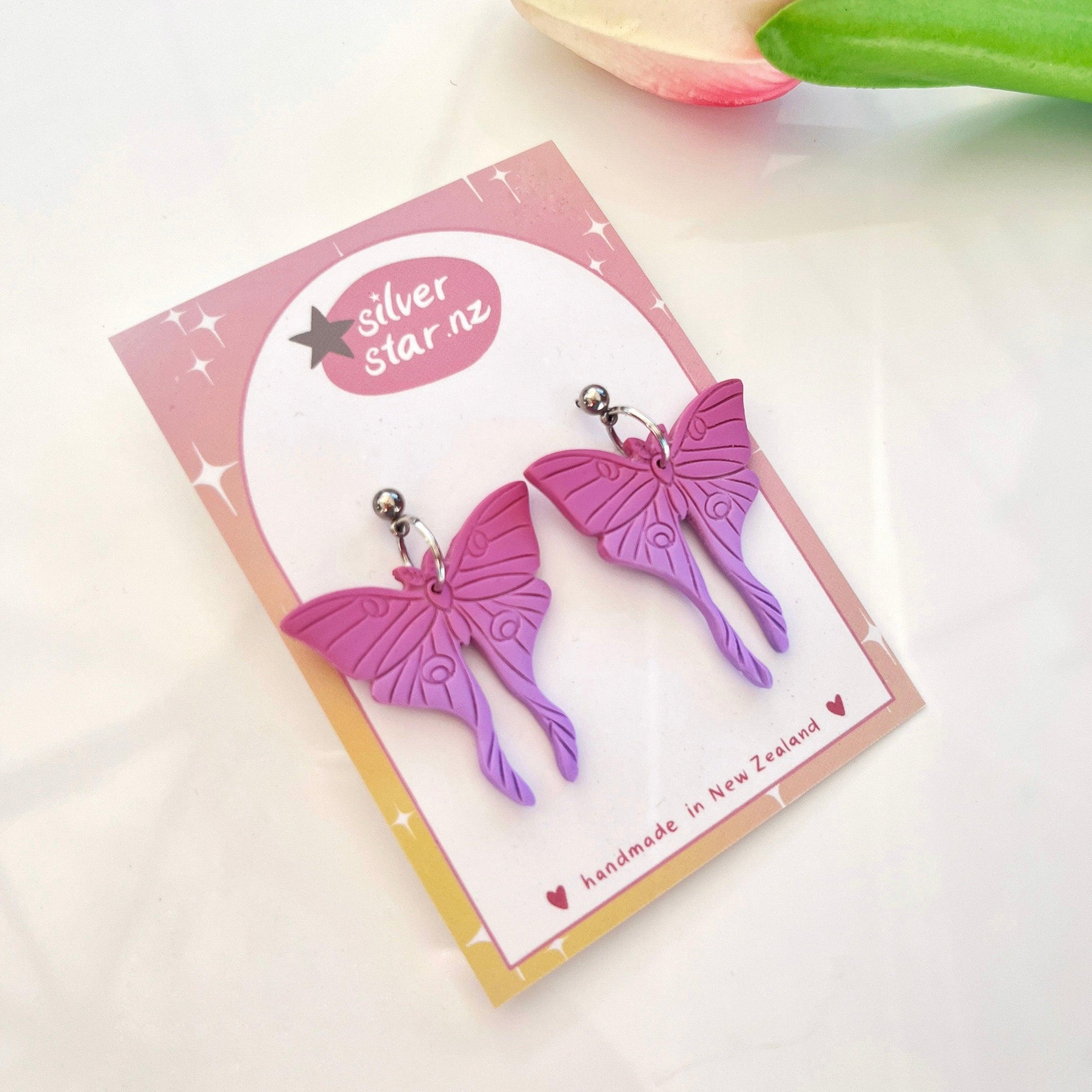 Moth Polymer Clay Earrings, resting on a pink card with silverstar.nz branding, are handmade in New Zealand and feature stainless steel hooks. Crafted from polymer clay, a tulip peeks from the corner, adding a touch of nature's charm.