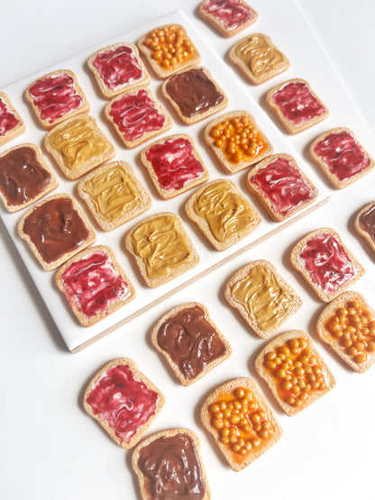 A collection of small toast-shaped cookies adorned with various toppings, including red jelly, chocolate, peanut butter, and orange jam. Much like the distinctive style found in silverstar.nz's Berry Jam on Toast Polymer Clay Earrings, these cookies are meticulously arranged on a white surface.