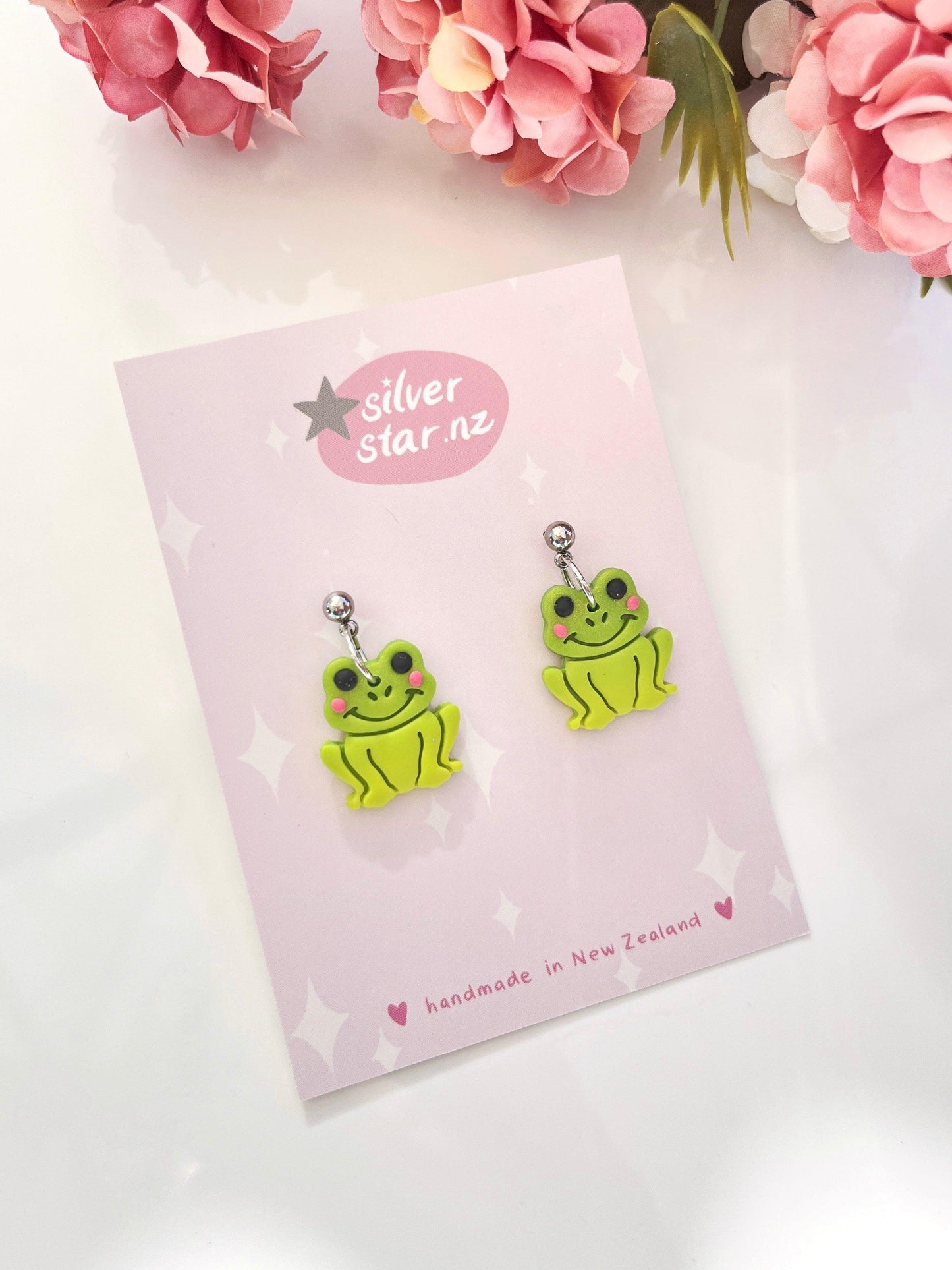 A pair of Frog Polymer Clay Earrings, featuring a charming design with green frogs and pink cheeks, is presented on a card that highlights the brand silver star.nz along with the text "handmade in New Zealand." These delightful handmade earrings include stainless steel hooks and are set against a backdrop adorned with pink springtime flowers.