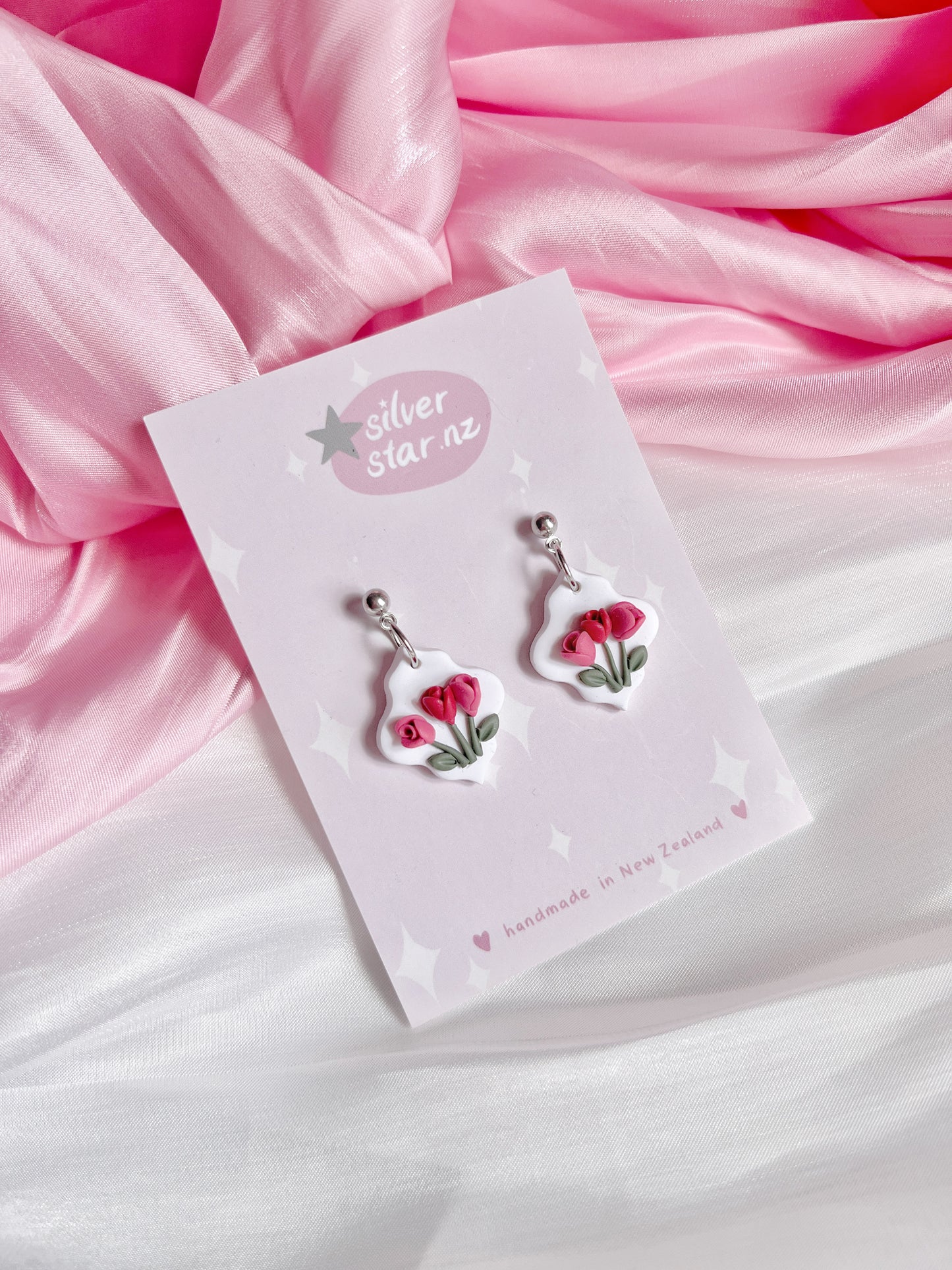 The Tulip Floral Polymer Clay Earrings, featuring pink flower pendants, are elegantly displayed on a card labeled silverstar.nz, resting on draped pink and white fabric. These silver earrings are handmade in New Zealand.