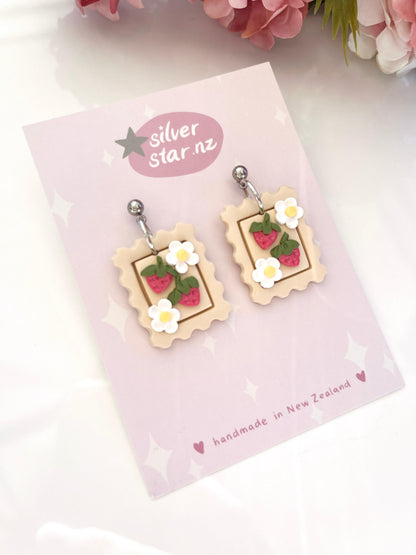 Introducing the Strawberry Stamp Polymer Clay Earrings, featuring postage stamp designs with strawberries and delicate white flowers. These handmade earrings are crafted from polymer clay and come elegantly mounted on a silver star.nz card. Equipped with stainless steel hooks, they're beautifully complemented by surrounding pink flower petals. Proudly made in New Zealand.