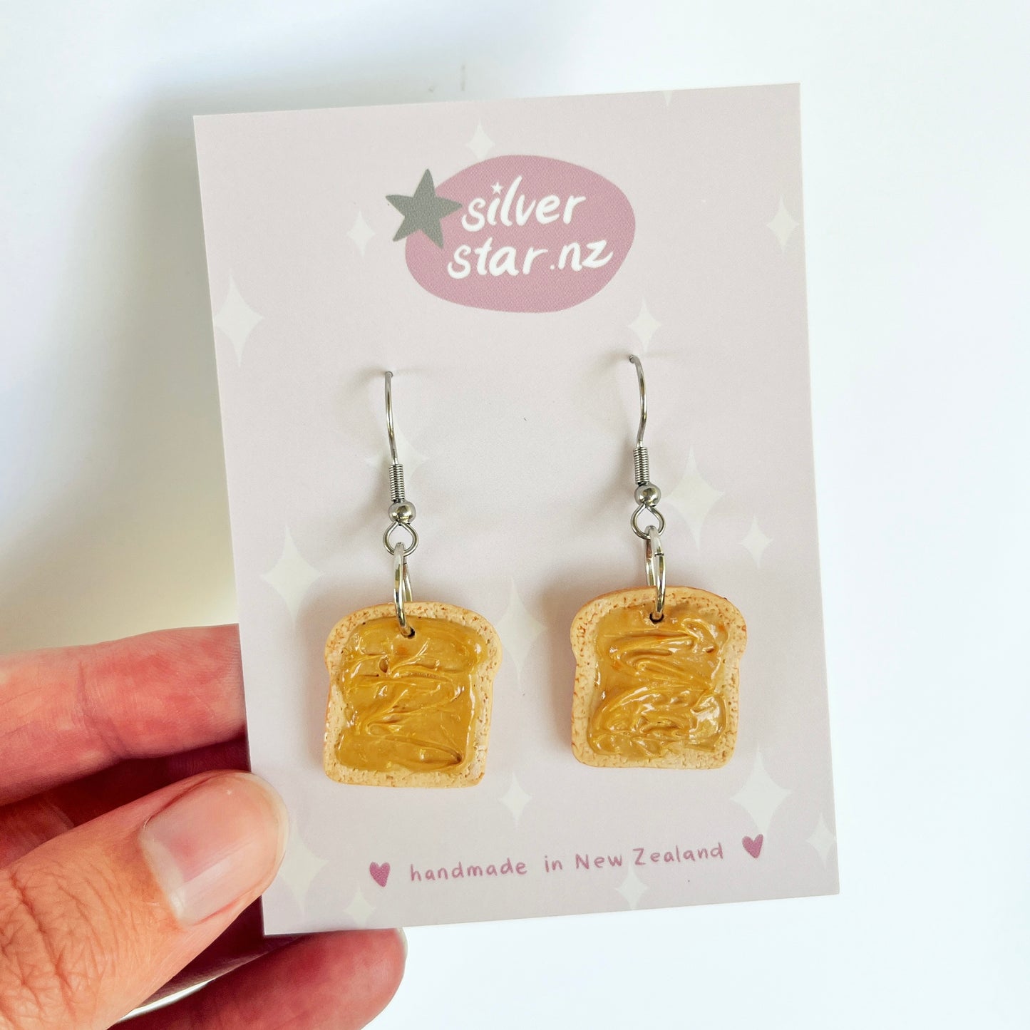 A hand displays the distinctively designed Peanut Butter on Toast Polymer Clay Earrings, resembling slices of bread with peanut butter. These earrings are crafted from handmade polymer clay and hang from stainless steel hooks, attached to a card featuring the silverstar.nz brand name, emphasizing their New Zealand origin. The background is plain white.