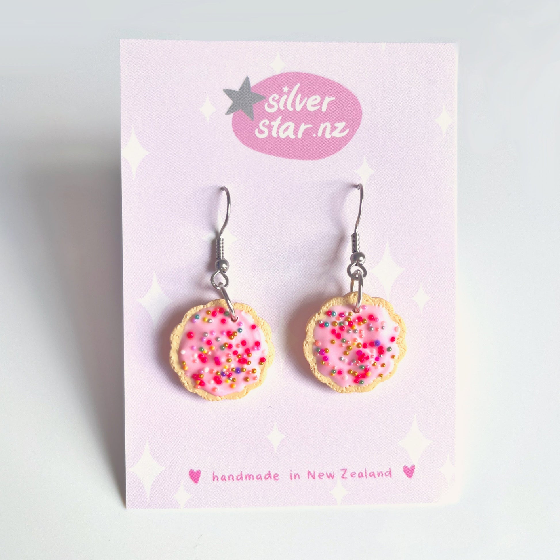 The Hundreds and Thousands Biscuit Earrings by silverstar.nz are crafted from polymer clay, resembling cookies with pink icing and colorful sprinkles. Displayed on a pink card labeled silverstar.nz, they feature stainless steel hooks, highlighting their handmade quality from New Zealand.