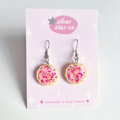 The Hundreds and Thousands Biscuit Earrings by silverstar.nz are crafted from polymer clay, resembling cookies with pink icing and colorful sprinkles. Displayed on a pink card labeled silverstar.nz, they feature stainless steel hooks, highlighting their handmade quality from New Zealand.