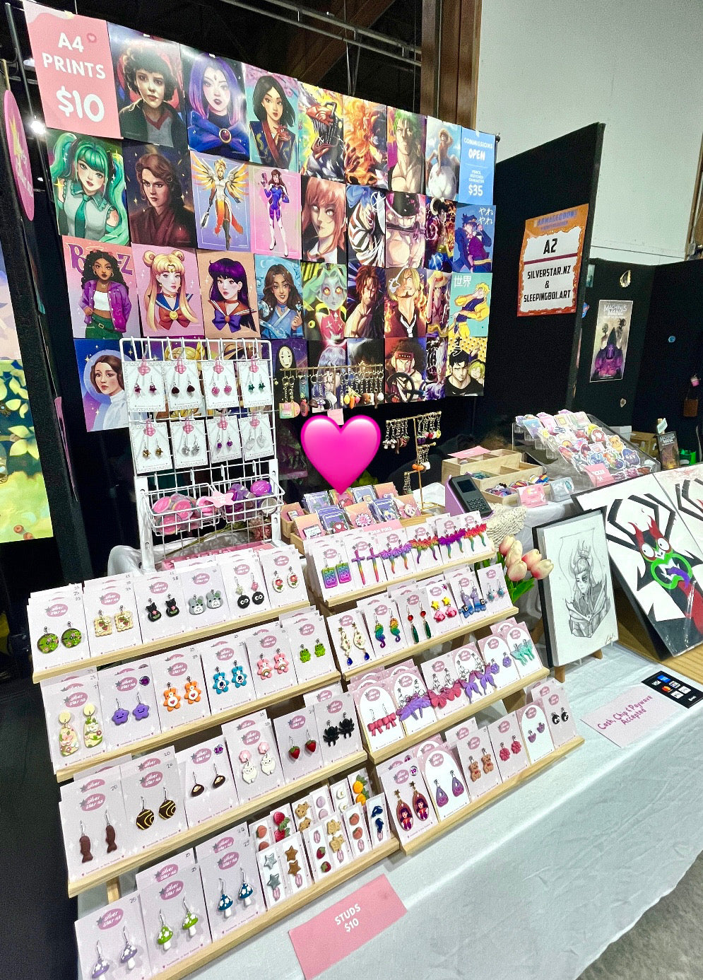 A vibrant vendor booth at a convention featuring colorful character art prints and a variety of jewelry. Displays include A4 prints priced at $10 and an array of earrings in different designs. A pink heart emoji covers part of the display.