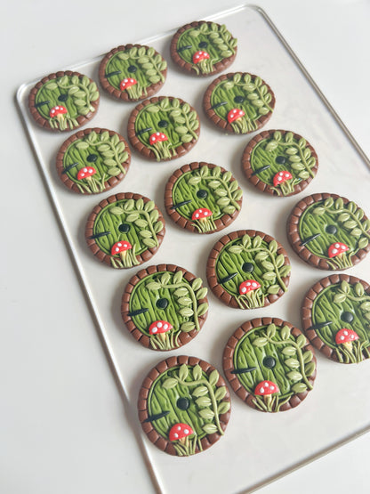 A set of Hobbit Door Polymer Clay Earrings by silverstar.nz, featuring round designs with green leafy motifs and red mushrooms, is elegantly displayed on a transparent tray against a white background.