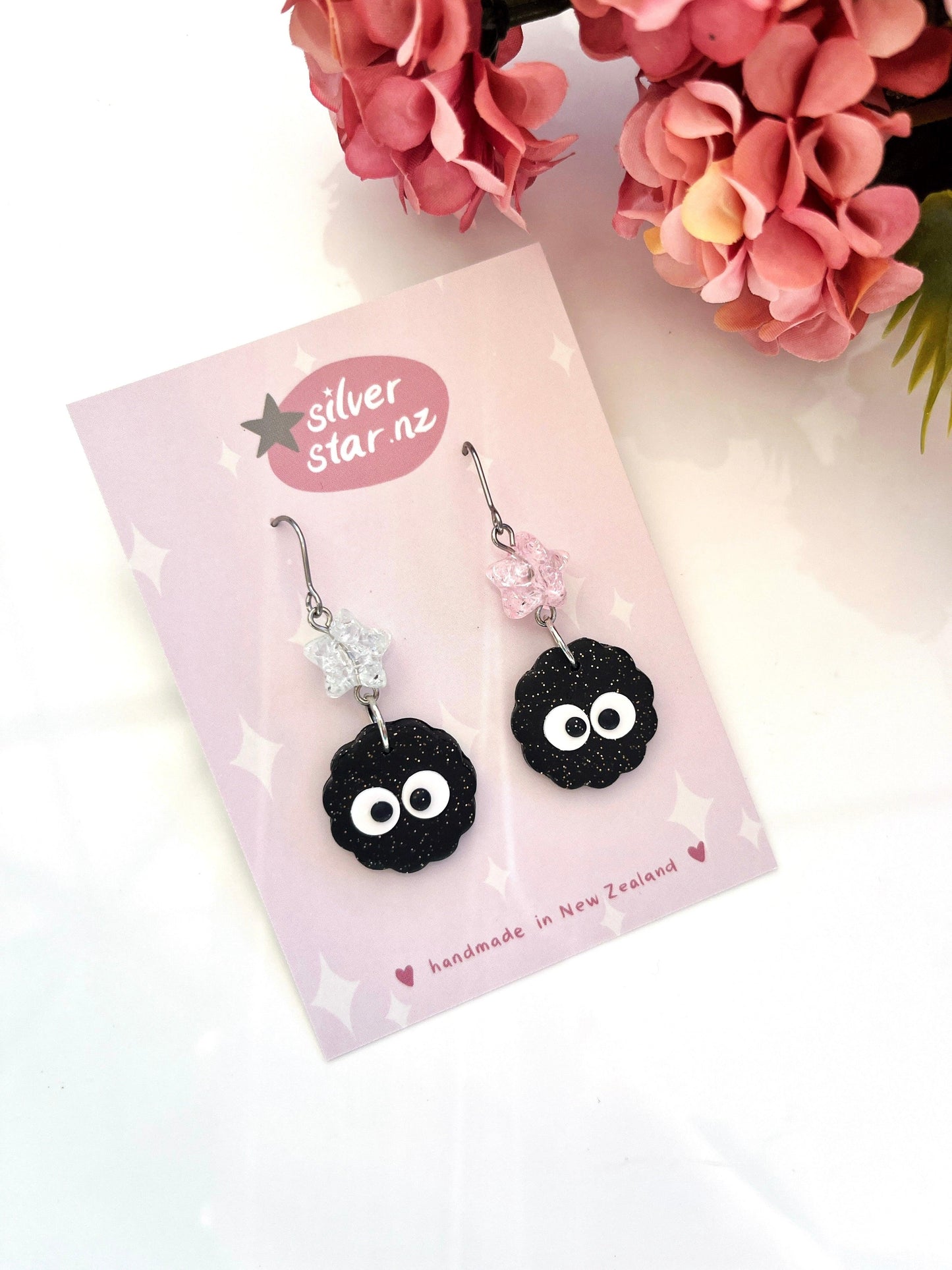 Displayed on a card with "silver star.nz" printed on it, the Soot Sprite Polymer Clay Earrings with Bead are handmade in New Zealand. The unique design features black, round characters with large white eyes attached to star-shaped hooks, accompanied by nearby pink flowers.