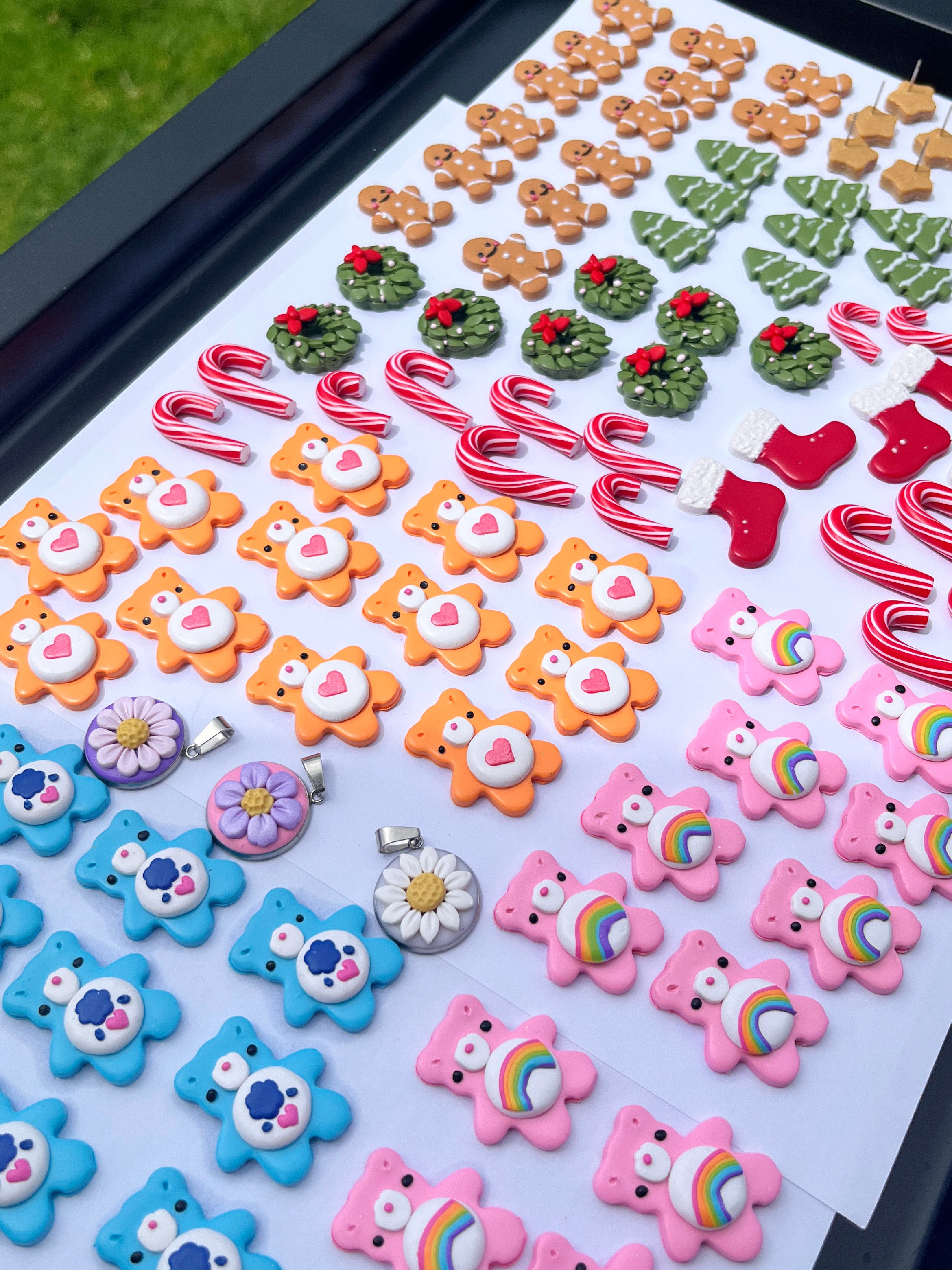 A display of colorful clay earrings shaped like teddy bears with hearts, candy canes, Christmas trees, stockings, and floral designs arranged neatly in rows on a board.