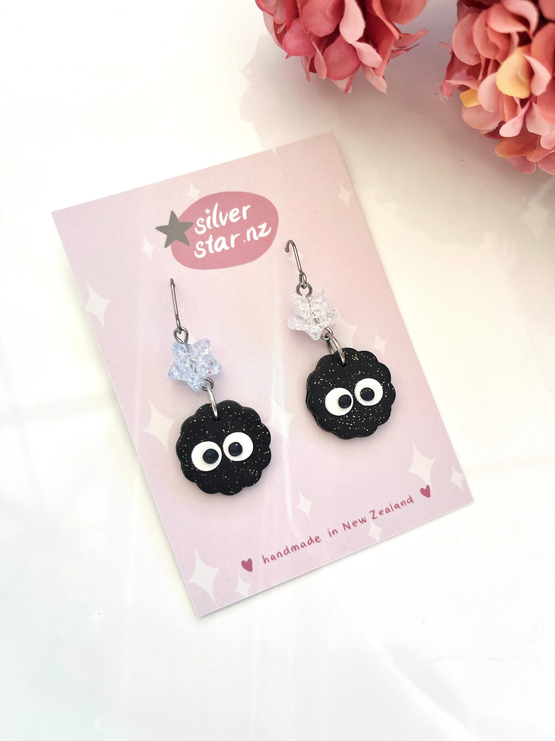 Pair of Soot Sprite Polymer Clay Earrings with Bead, featuring handmade black soot sprites with white eyes hanging from silver hooks. Each earring includes a translucent star bead above the sprite. Presented on a pink card that reads "silver star.nz" and "unique design from New Zealand.