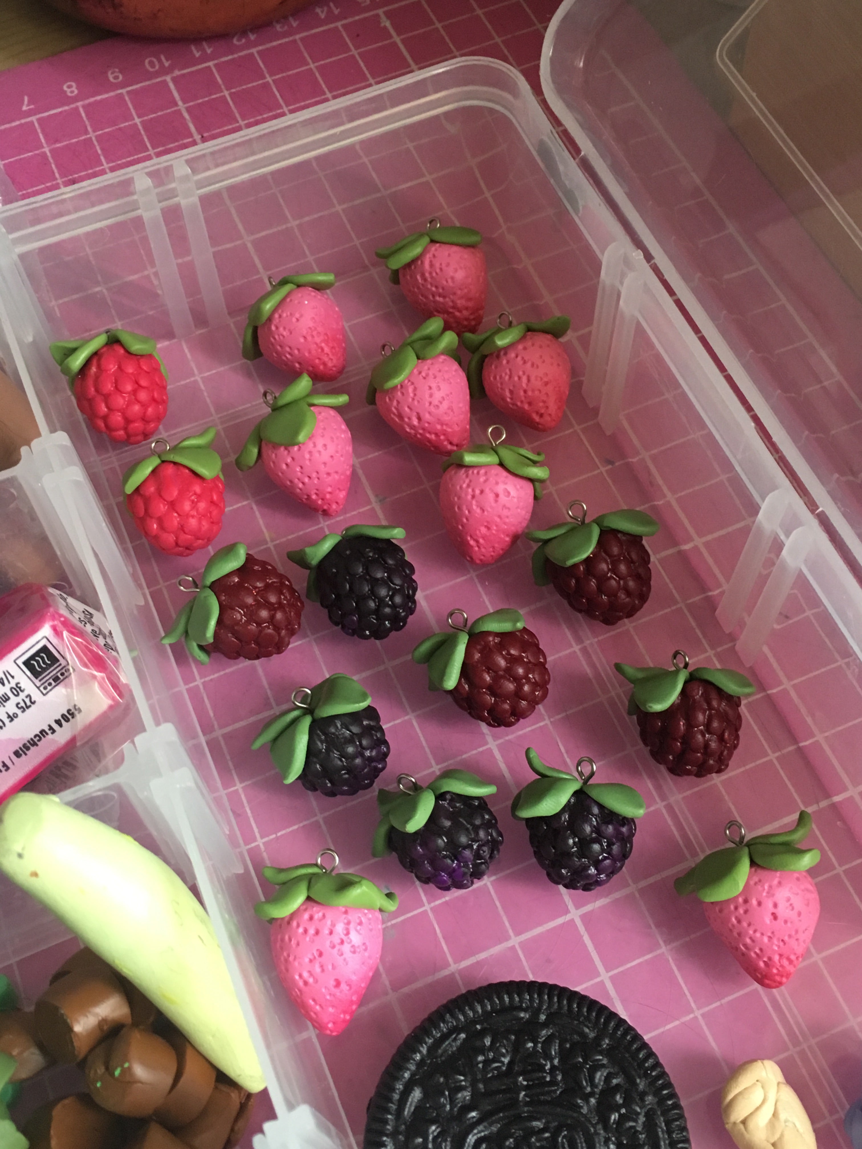 A collection of handmade polymer clay charms shaped like strawberries, raspberries, blackberries, and an Oreo cookie are displayed in a clear plastic organizer on a pink grid-patterned surface.