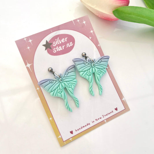 A pair of mint green Moth Polymer Clay Earrings with intricate designs is displayed on a card. The card reads "silver star.nz," indicating they are handmade in New Zealand. Crafted from polymer clay and featuring stainless steel hooks, a pink tulip is partially visible in the background.