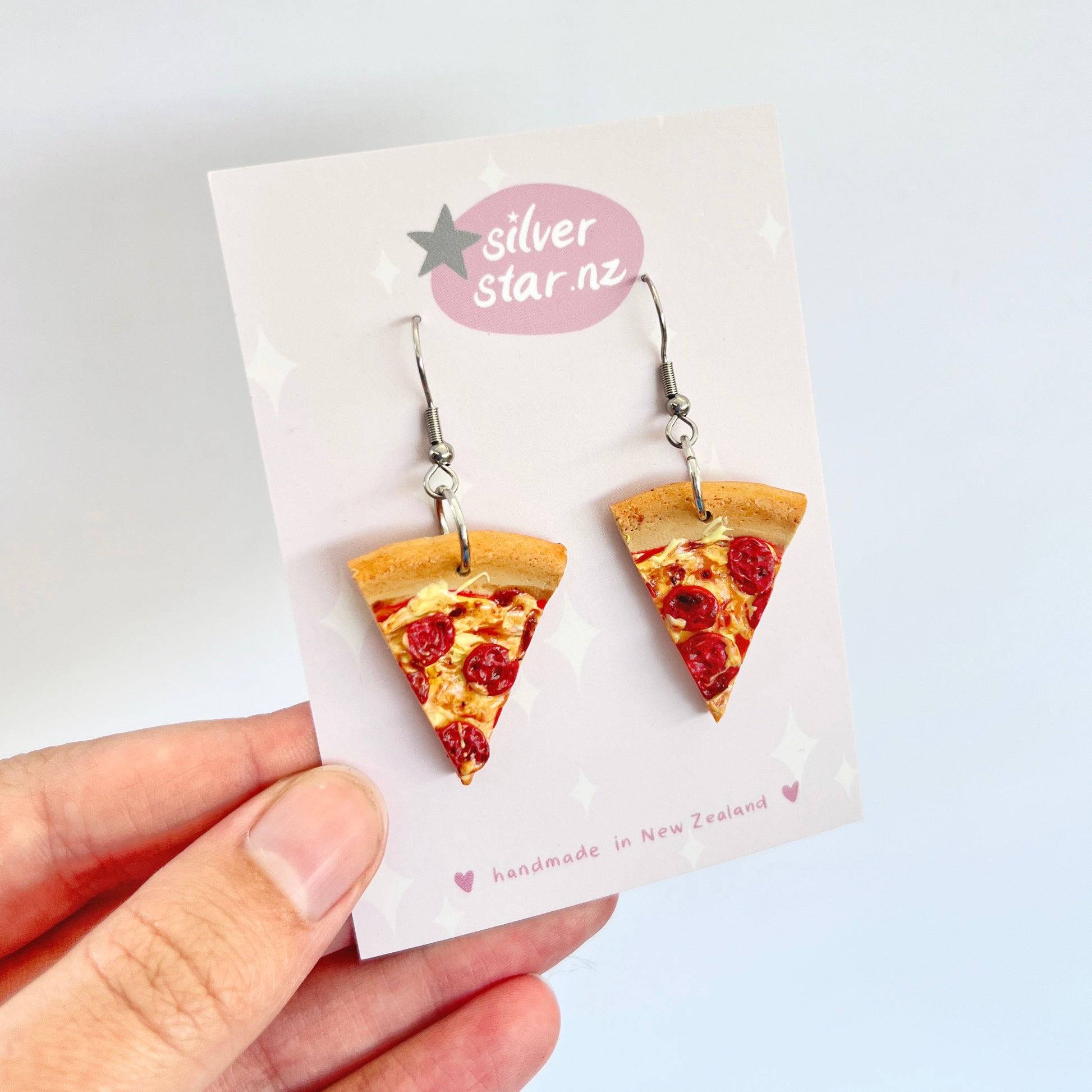 A hand showcases a pair of pizza slice-shaped novelty earrings with pepperoni toppings, crafted from polymer clay. These handmade "Pizza Polymer Clay Earrings" dangle from stainless steel hooks and are presented on a card bearing the silverstar.nz logo with the text "handmade in New Zealand.