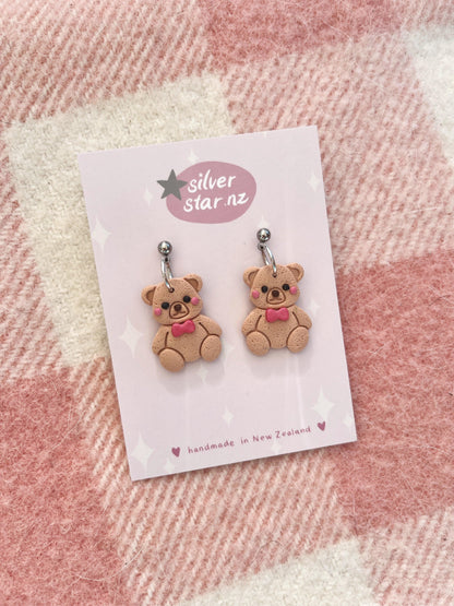 A pair of Teddy Polymer Clay Earrings, showcasing a delightful design of whimsical bears adorned with pink bows, is artfully attached to a card labeled silver star.nz. These earrings feature a distinctive funky style and are expertly crafted in New Zealand with stainless steel hooks. They are beautifully presented against a pink and white checkered fabric background.