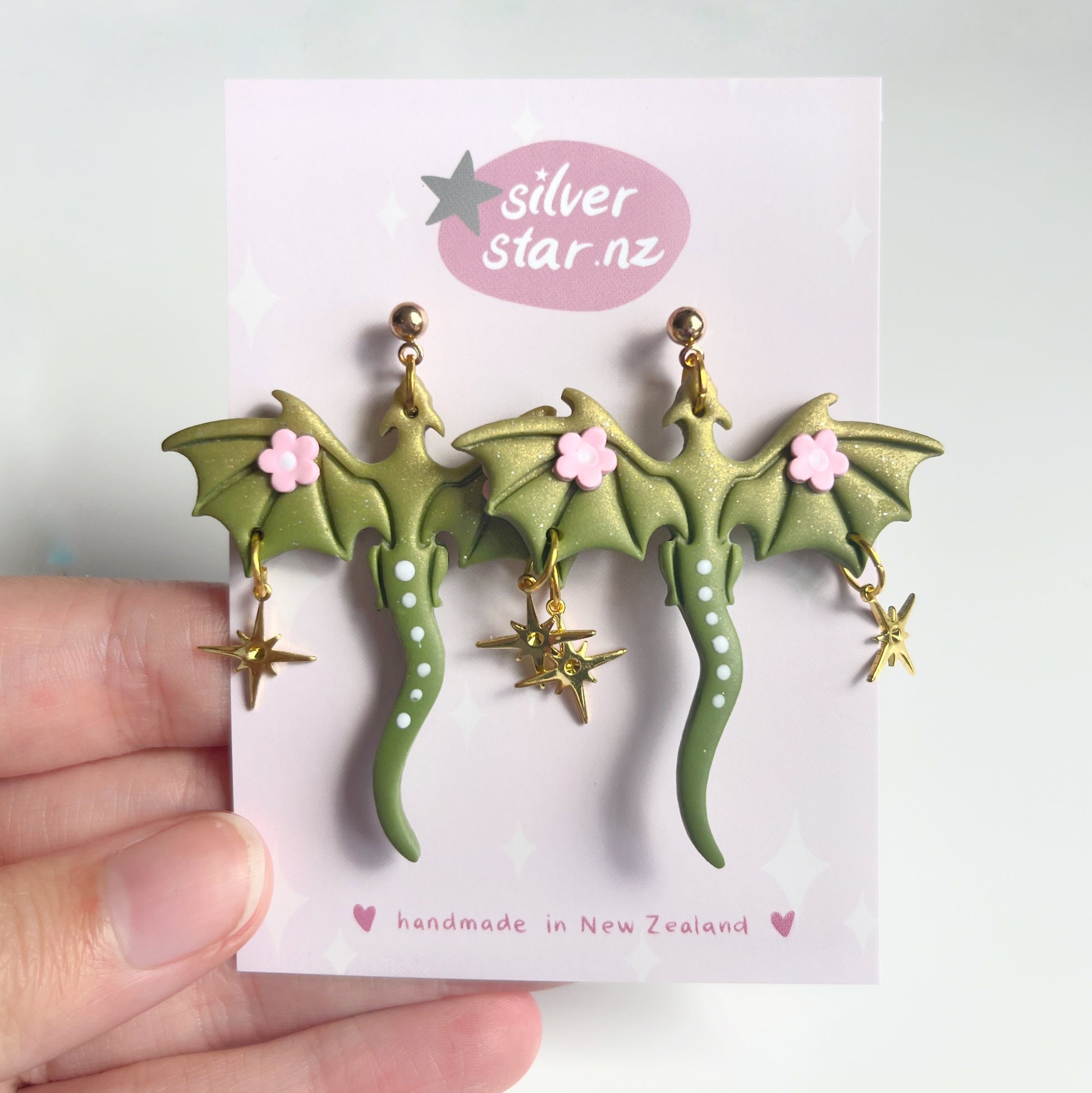 A hand displays a card featuring Green Fae Dragon Polymer Clay Earrings, adorned with wings, pink flowers, and star charms. The card reads silverstar.nz and handmade in New Zealand, emphasizing their artisanal charm.