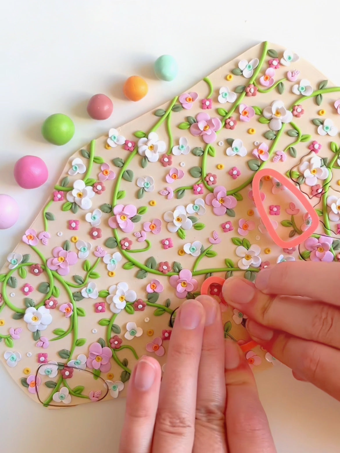 Hands are crafting with colorful polymer clay, pressing flower and leaf patterns onto a slab using tools. Round balls of clay in various pastel colors are visible, showcasing a delicate and intricate floral design.