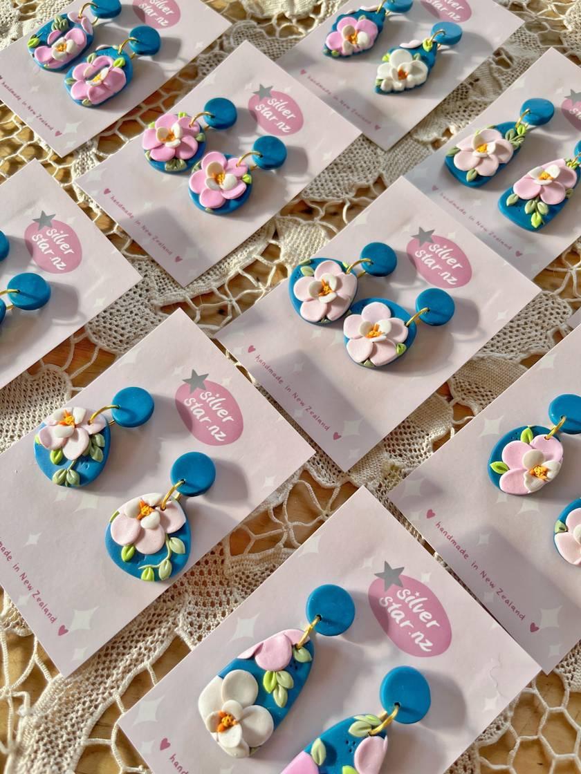 Colorful earrings with floral designs displayed on cards labeled Silver Star. The earrings include blue bases adorned with pink and white flowers. They are arranged on a textured lace surface.