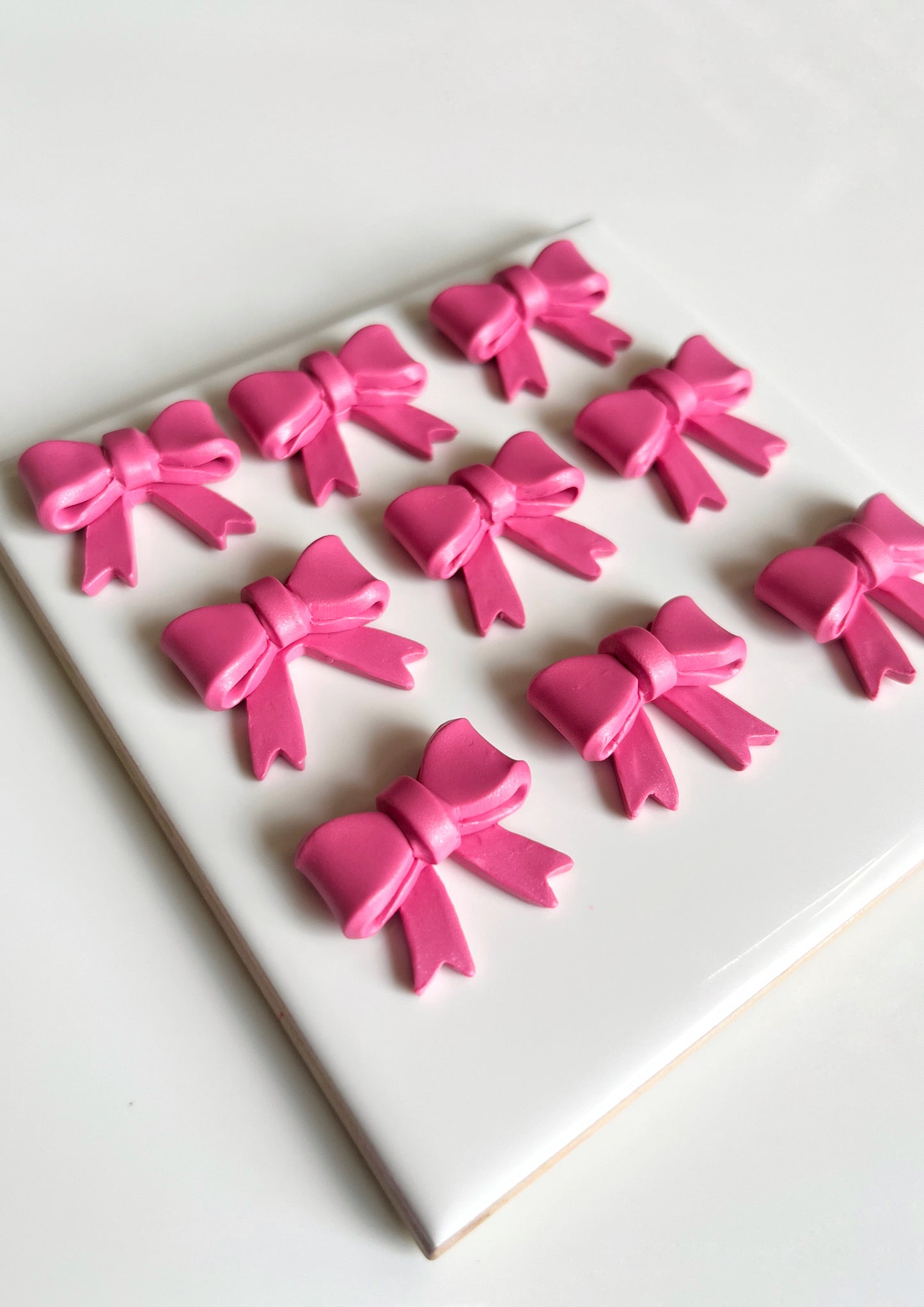 The image features Bow Coquette Polymer Clay Earrings from silverstar.nz, beautifully arranged on a white ceramic tile with nine pink ribbon bows aligned in a 3x3 grid, creating a charming symmetrical pattern against the pristine background.