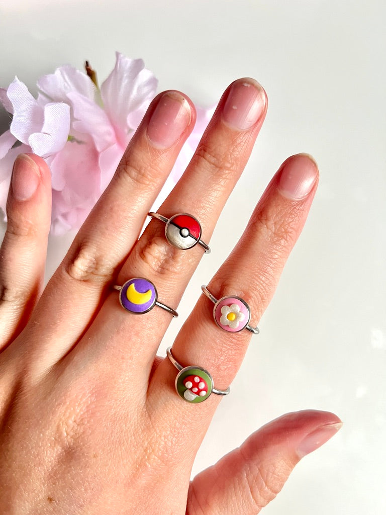 A hand displaying four colorful rings, each with a unique design: a red and white ball, a crescent moon, a flower, and a mushroom. A light pink flower is seen in the background.