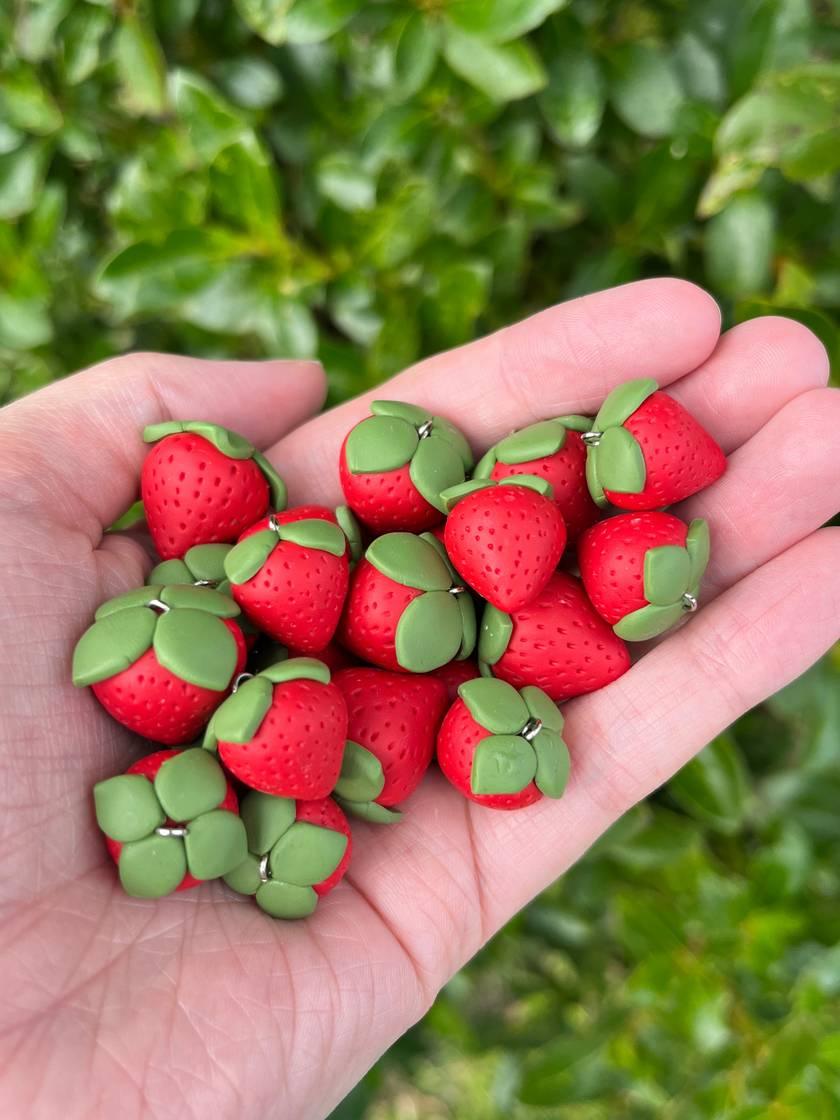 A hand holding small, handcrafted clay strawberries with green leaves. The strawberries are vibrant red with textured surfaces, resembling real fruit, set against a blurred leafy background.