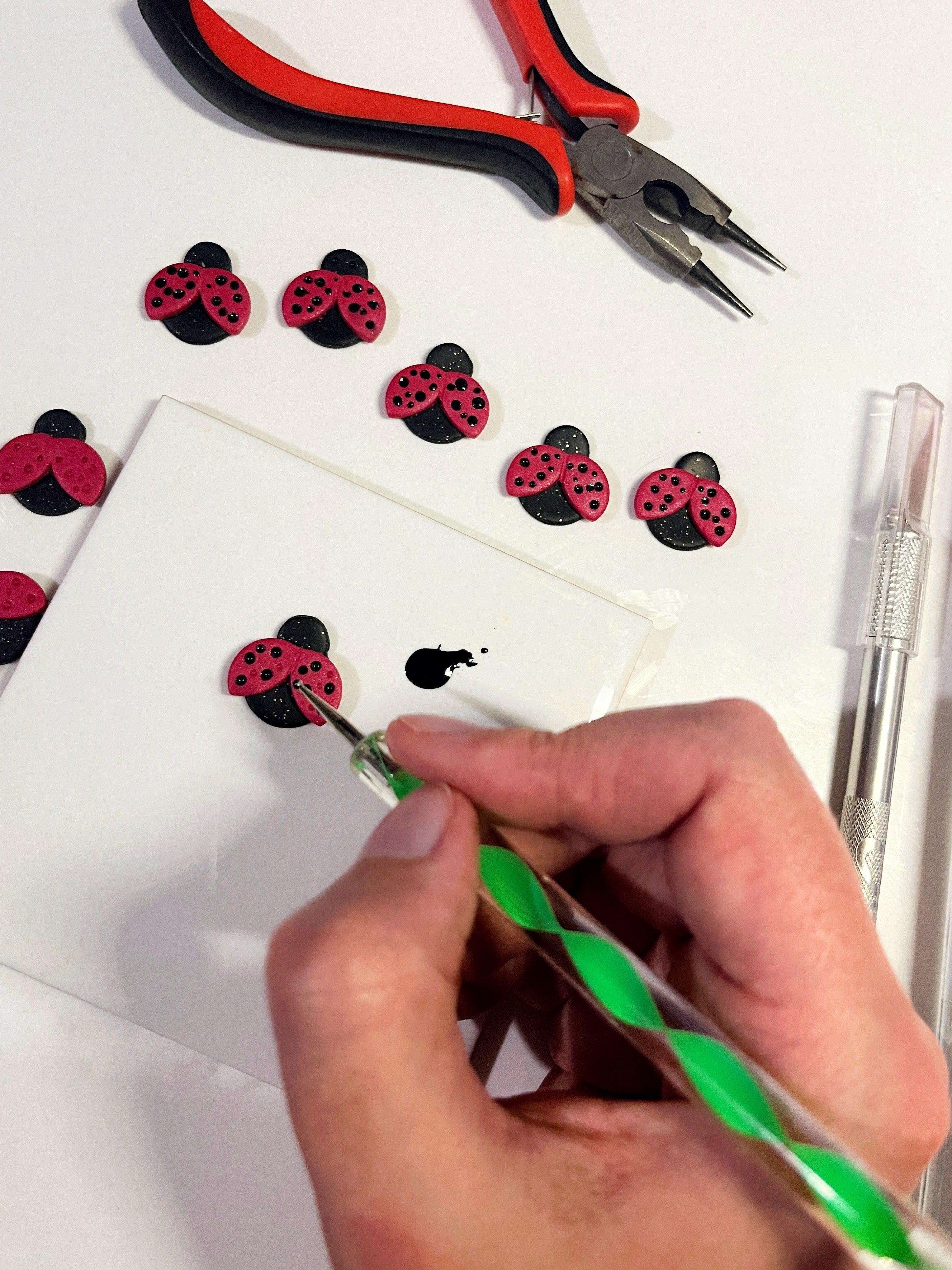 A person uses a tool to add black paint details to red and black ladybug-shaped crafts on a white tile. Several finished ladybugs are lined up, and pliers, a craft knife, and a green stylus are visible nearby.