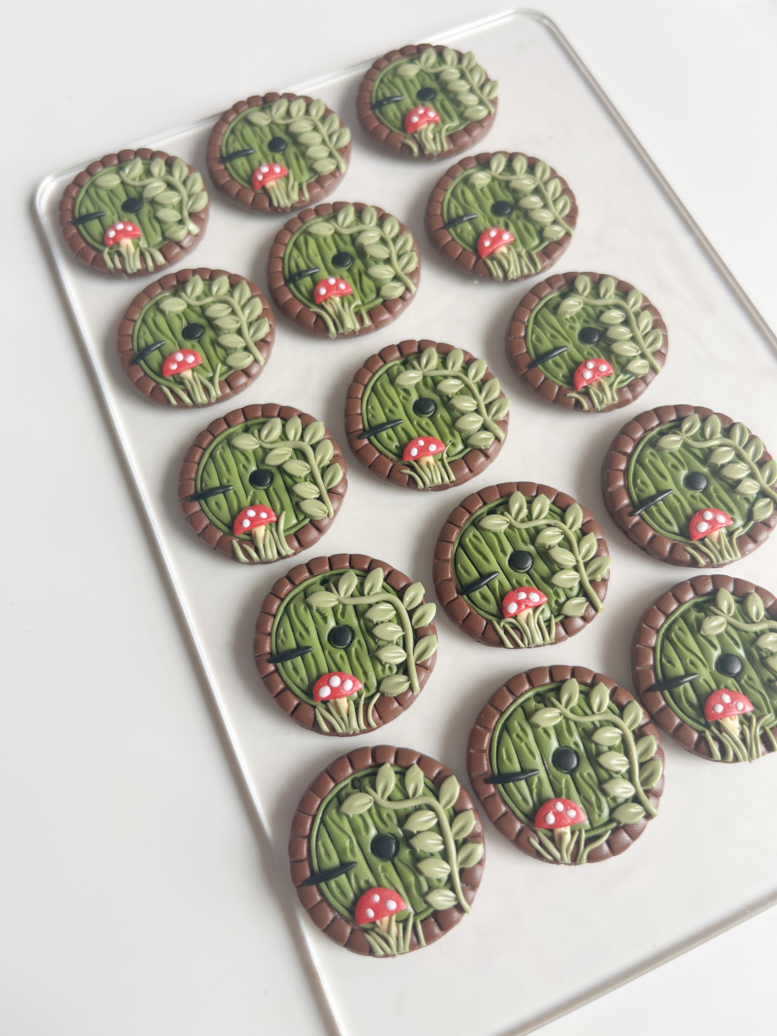 A set of Hobbit Door Polymer Clay Earrings by silverstar.nz, featuring round designs with green leafy motifs and red mushrooms, is elegantly displayed on a transparent tray against a white background.