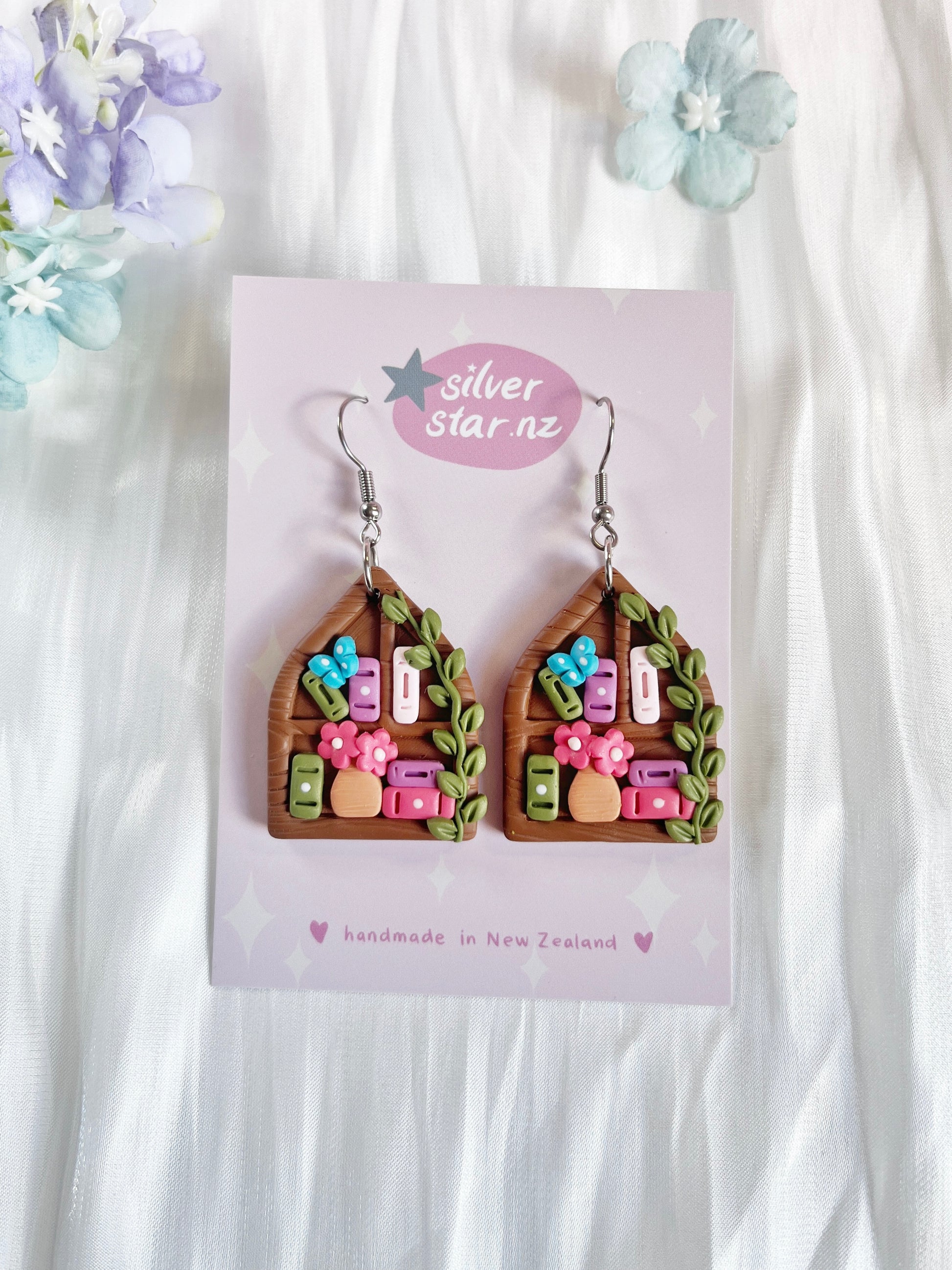 Pointed Arch Bookshelf Polymer Clay Earrings by silverstar.nz feature brown house shapes with pink flowers, greenery, and colorful rectangles on stainless steel hooks. Displayed on a silverstar.nz card against a white floral background.