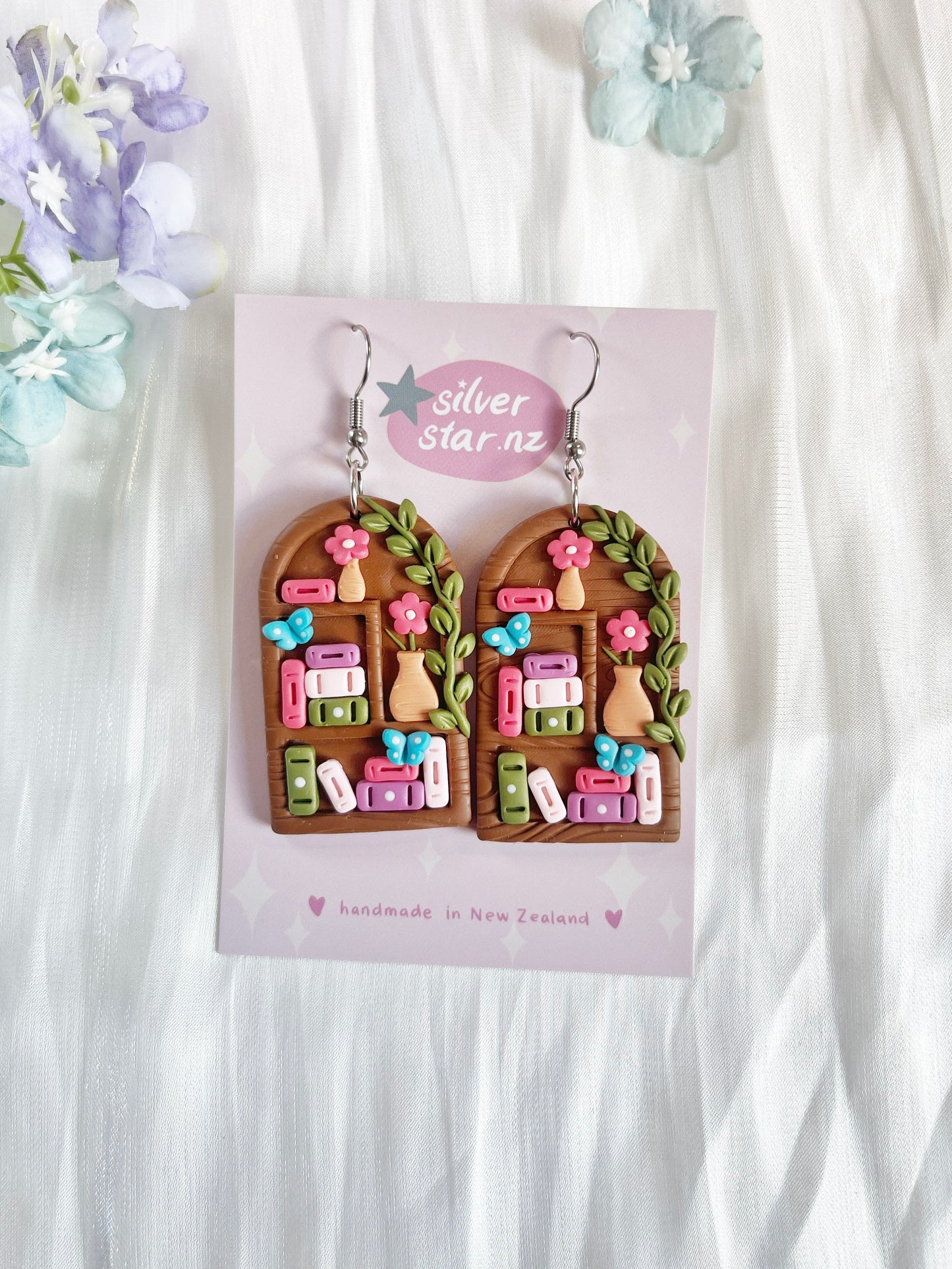 Tall Arch Bookshelf Polymer Clay Earrings by silverstar.nz feature colorful books, potted plants, and pink flowers on arched shapes with stainless steel hooks. Presented on a branded card with a floral background.