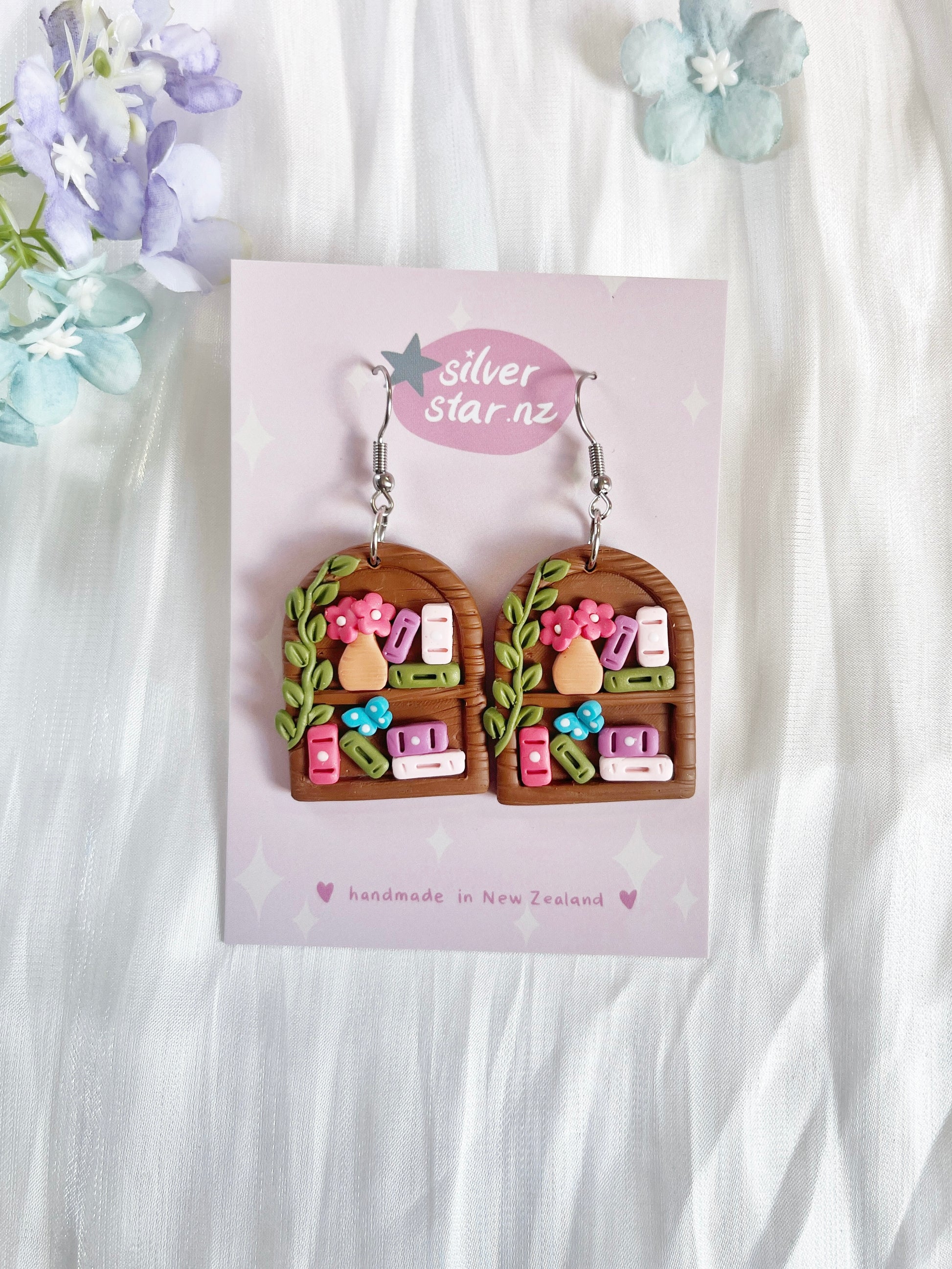 Round Arch Bookshelf Polymer Clay Earrings by silverstar.nz, handmade and featuring arched window shapes with colorful books, plants, and flowers. Displayed on a pastel silverstar.nz card with delicate floral decor in the background.