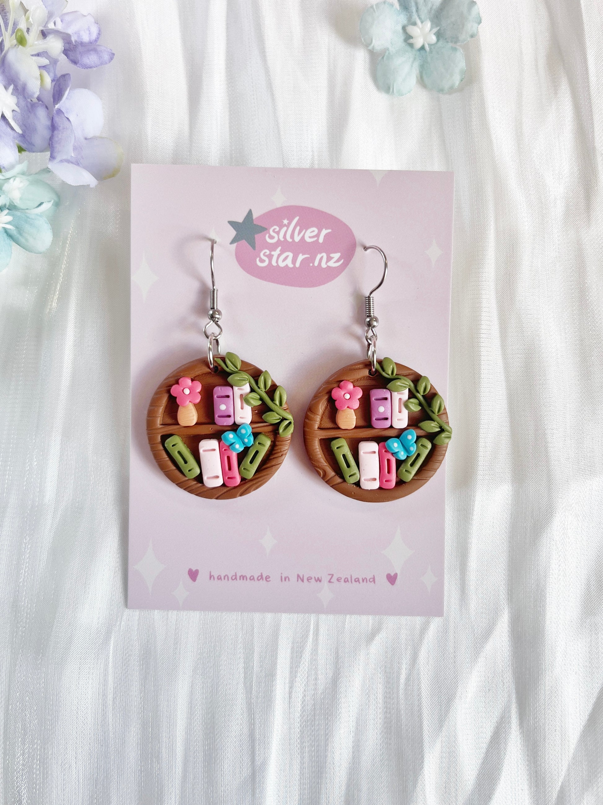Round Bookshelf Polymer Clay Earrings by silverstar.nz—handmade, round earrings featuring colorful miniature books, flowers, and leaves. Displayed on a branded card with pastel stars and hearts for a unique look.