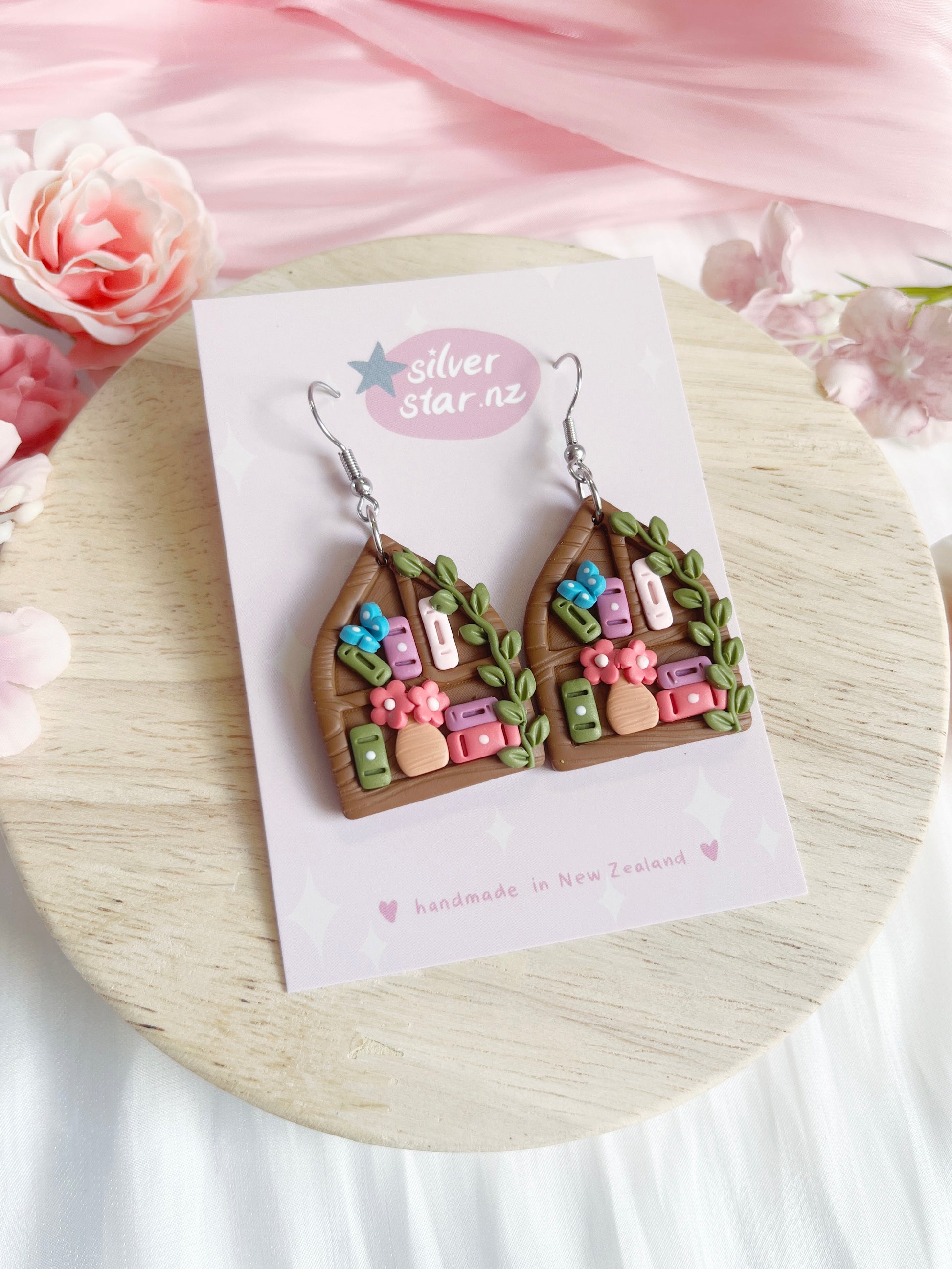 Pointed Arch Bookshelf Polymer Clay Earrings by silverstar.nz, displayed on a branded card with pink flowers on a light wooden surface. Features colorful details and stainless steel hooks.